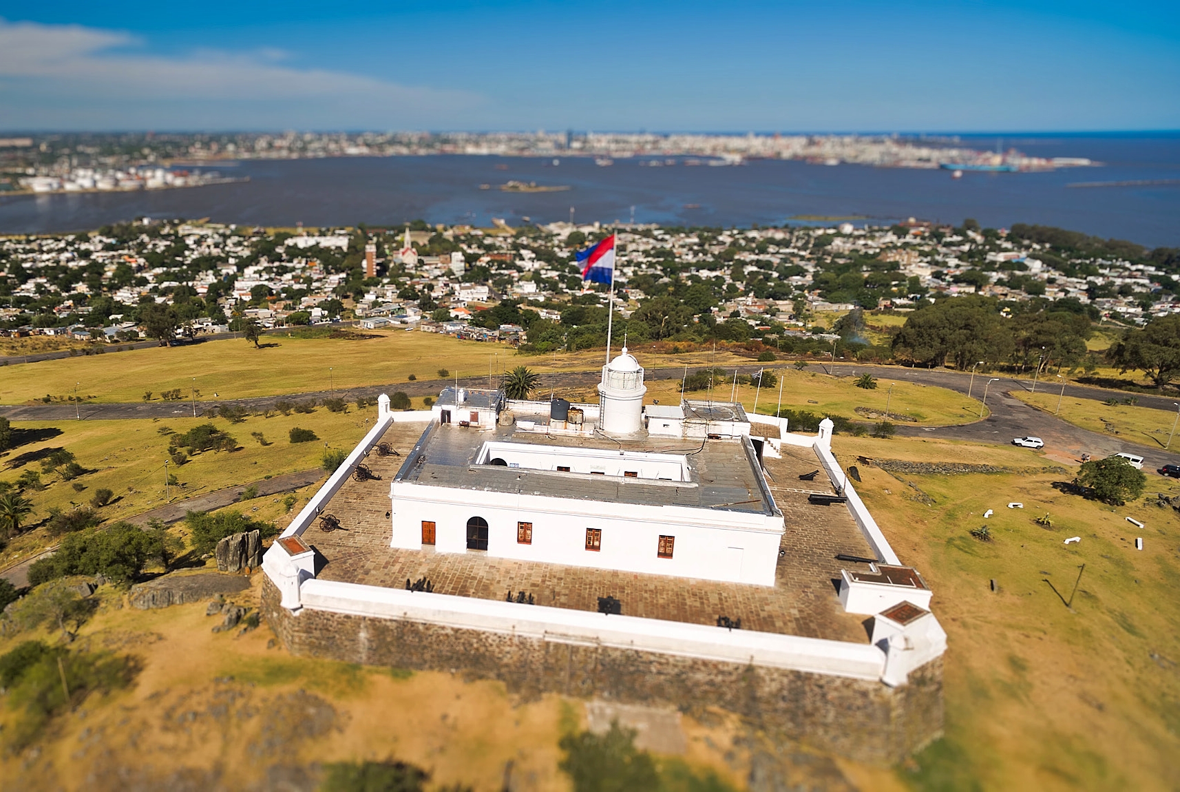 500px provided description: Military Museum fortress Hill, bay view Montevideo Uruguay [#sky ,#forest ,#people ,#blue ,#wind ,#clouds ,#cars ,#cityscape ,#tilt-shift ,#beautiful ,#green ,#fun ,#hill ,#bay ,#flag ,#urban exploration ,#Fortress]