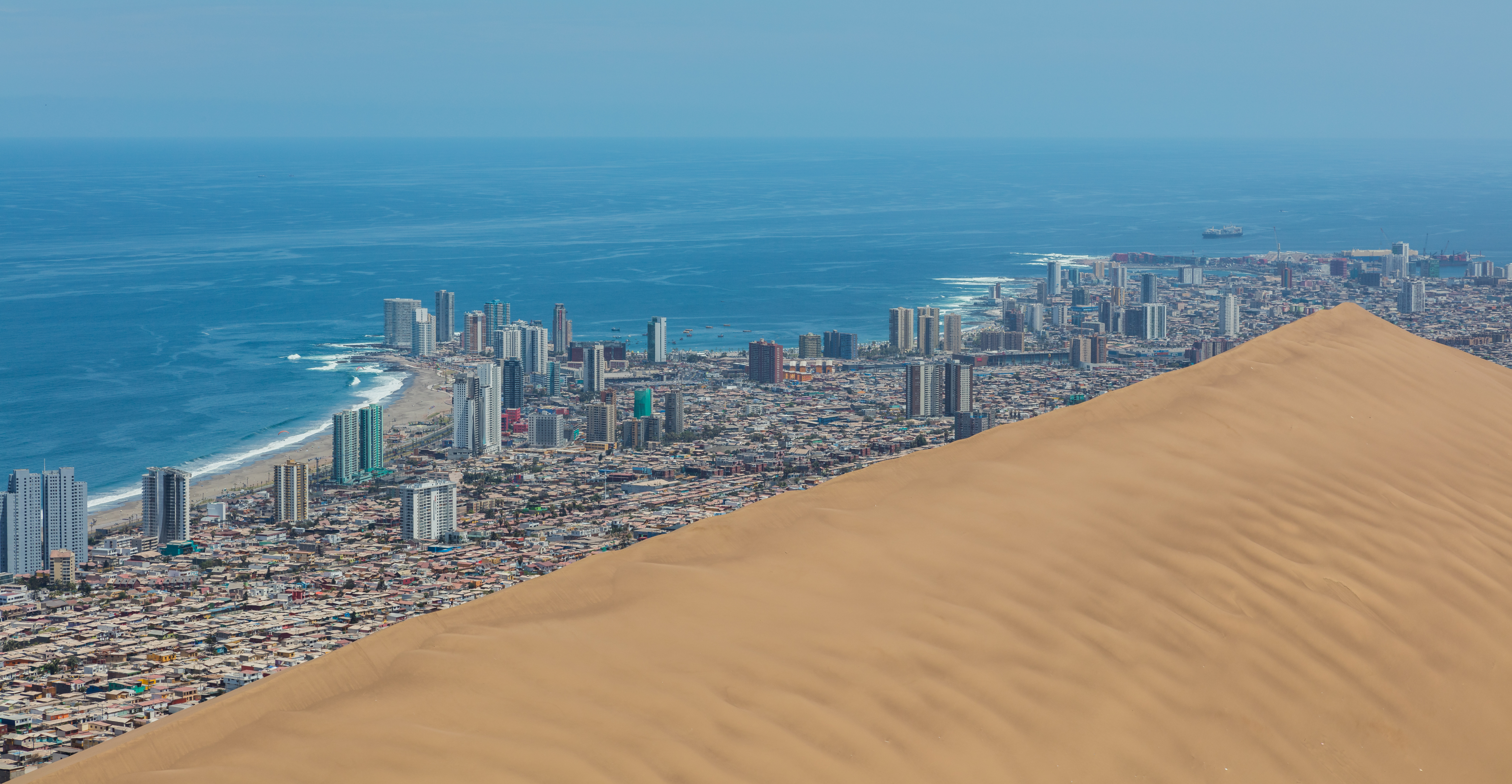 View of the Dragon Dune in the foreground and the city of Iquique in the background, Tarapacá Region, northern Chile. The dune is about 20 000 years old and was originated by coastal winds when the sea level was 100 metres (110 yd) further inside. The dune looks menacing, especially from the bottom, but is stable. It became a Natural Sanctuary in 2005 but before that it was partially removed in the West wing to allow the growth of the city of Iquique. The dune is 6.4 kilometres (4.0 mi) long, between 150–550 metres (160–600 yd) wide and 320 metres (350 yd) high. Iquique has aprox. 185 000 inhabitants and is a prosperous and fast-growing city thanks to the free trade activities.