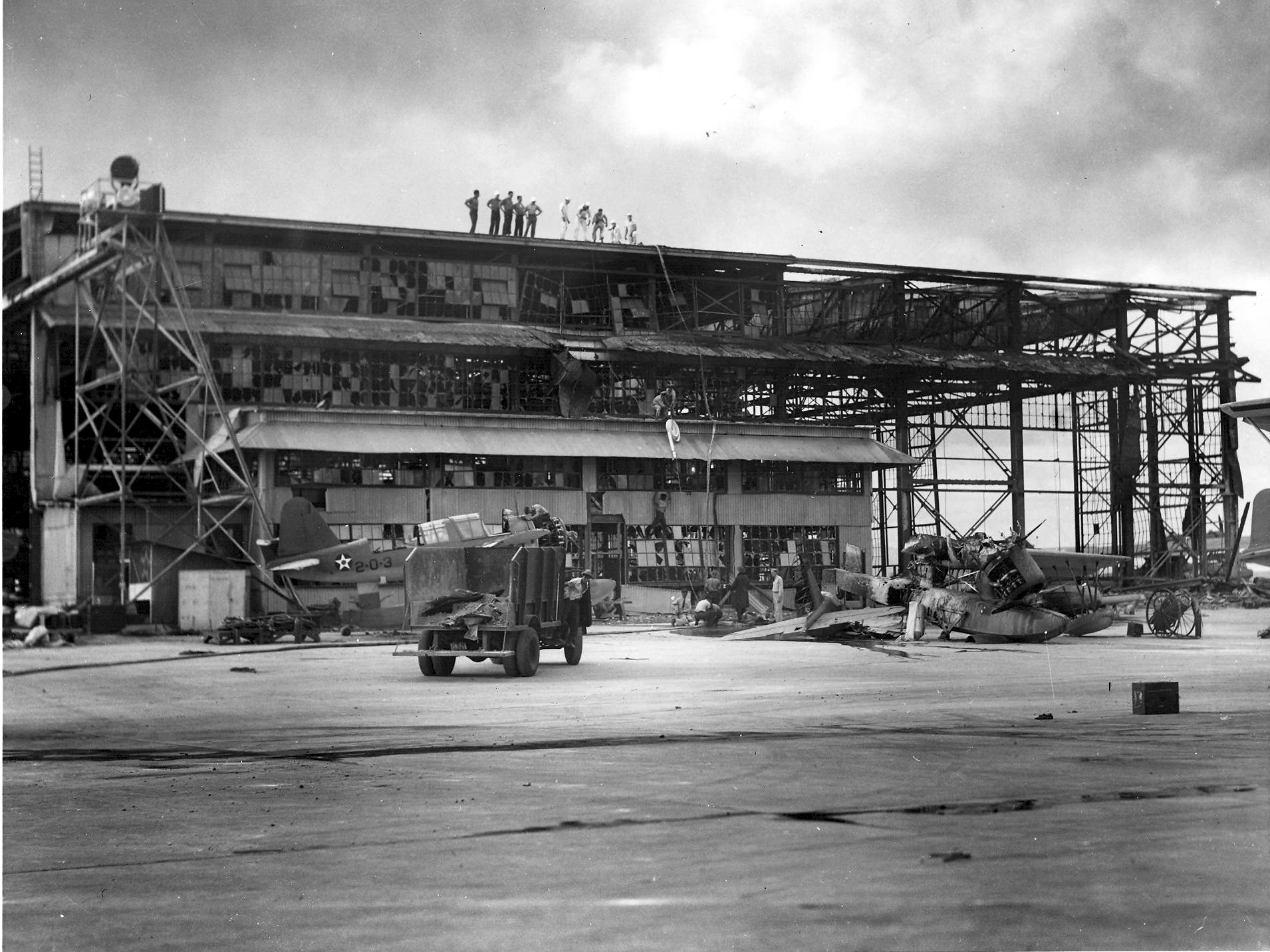 U.S. Navy personnel inspect a damaged hangar at Naval Air Station Ford Island in the aftermath of the Japanese attack on Pearl Harbor, Hawaii (USA), December 1941. A destroyed Vought OS2U Kingfisher is visible on the right, another OS2U of Observation Squadron VO-2 is worked on.