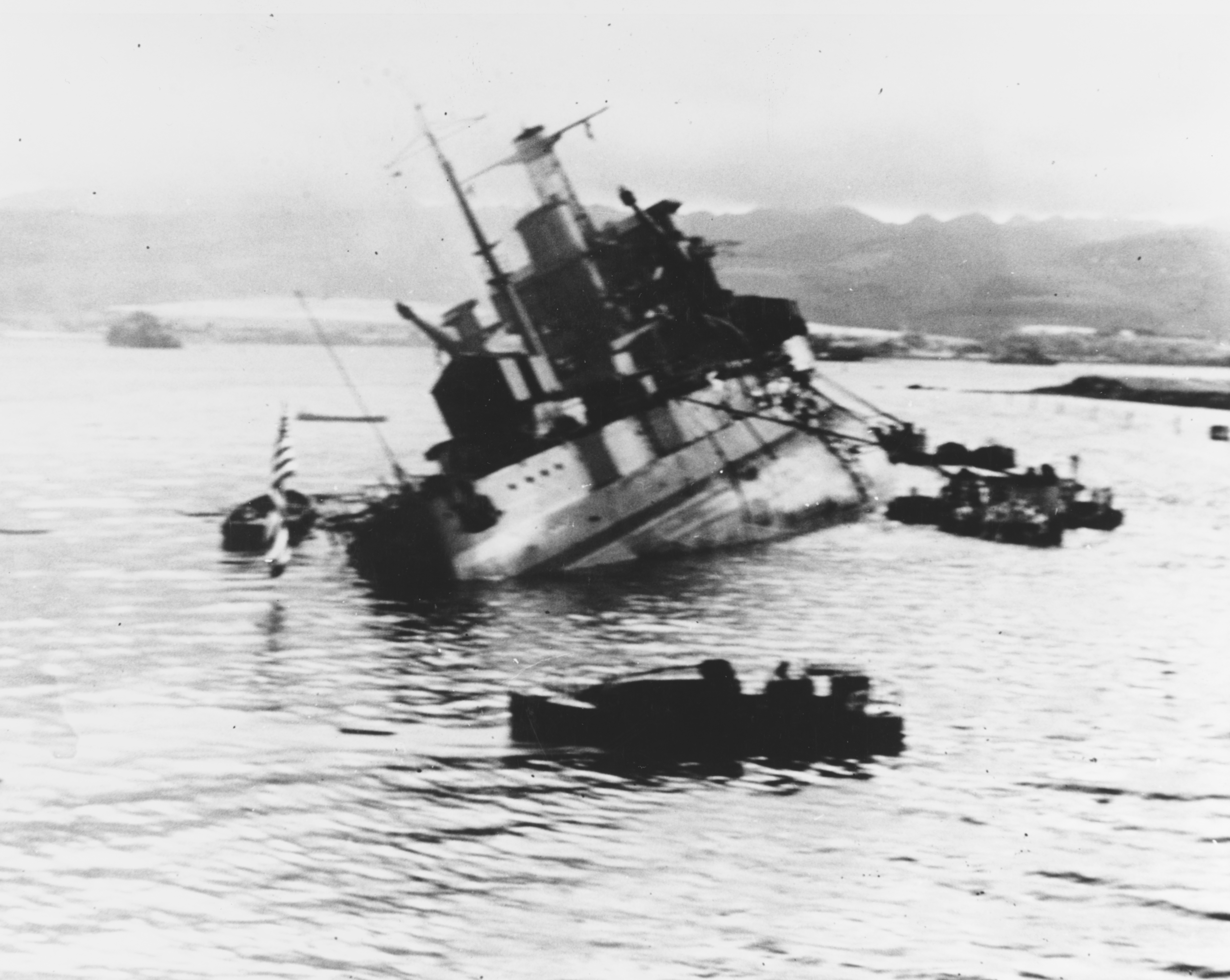 The U.S. Navy target ship USS Utah (AG-16, ex BB-31) capsizing off Ford Island, during the attack on Pearl Harbor, Hawaii (USA), 7 December 1941, after being torpedoed by Japanese aircraft. Photographed from USS Tangier (AV-8), which was moored astern of Utah. Note colours half-raised over fantail, boats nearby, and sheds covering Utah´s after guns.