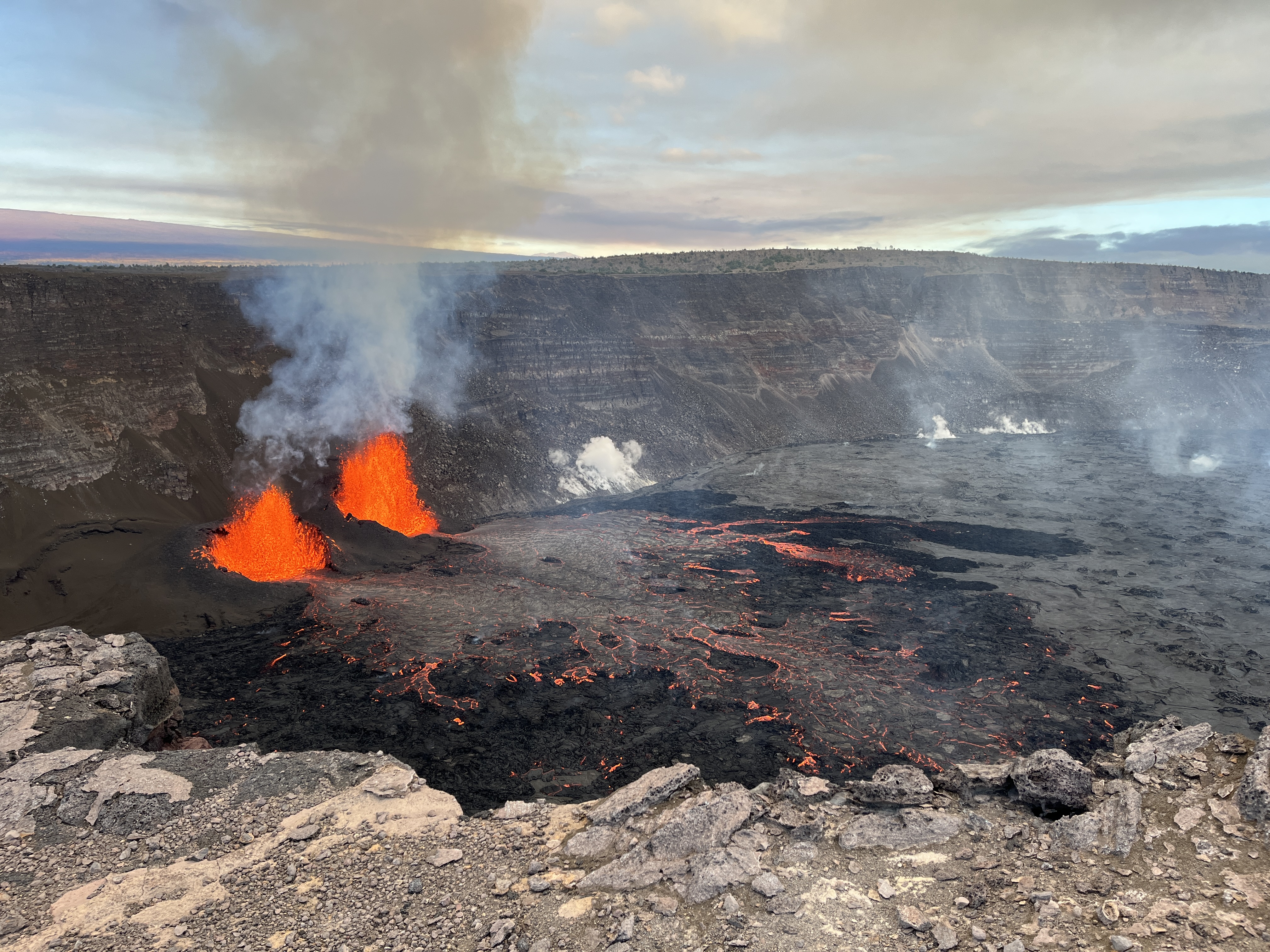 View of the Kīlauea summit eruption in Hawaiʻi Volcanoes National Park, from the south rim of the caldera. The caldera wall behind the erupting vents is approximately 210 meters (690 feet) tall. This morning, USGS Hawaiian Volcano Observatory field crews reported lava fountain heights of 50 meters (160 feet) and 40 m (140 feet) from north and south vents, respectively. At about 8:25 a.m. this morning, field crews reported that parts of the south vent collapsed, with an associated reduction of about half in south vent lava fountain height. Lava flow activity has remained confined to the southwest crater floor area near the active vents.
