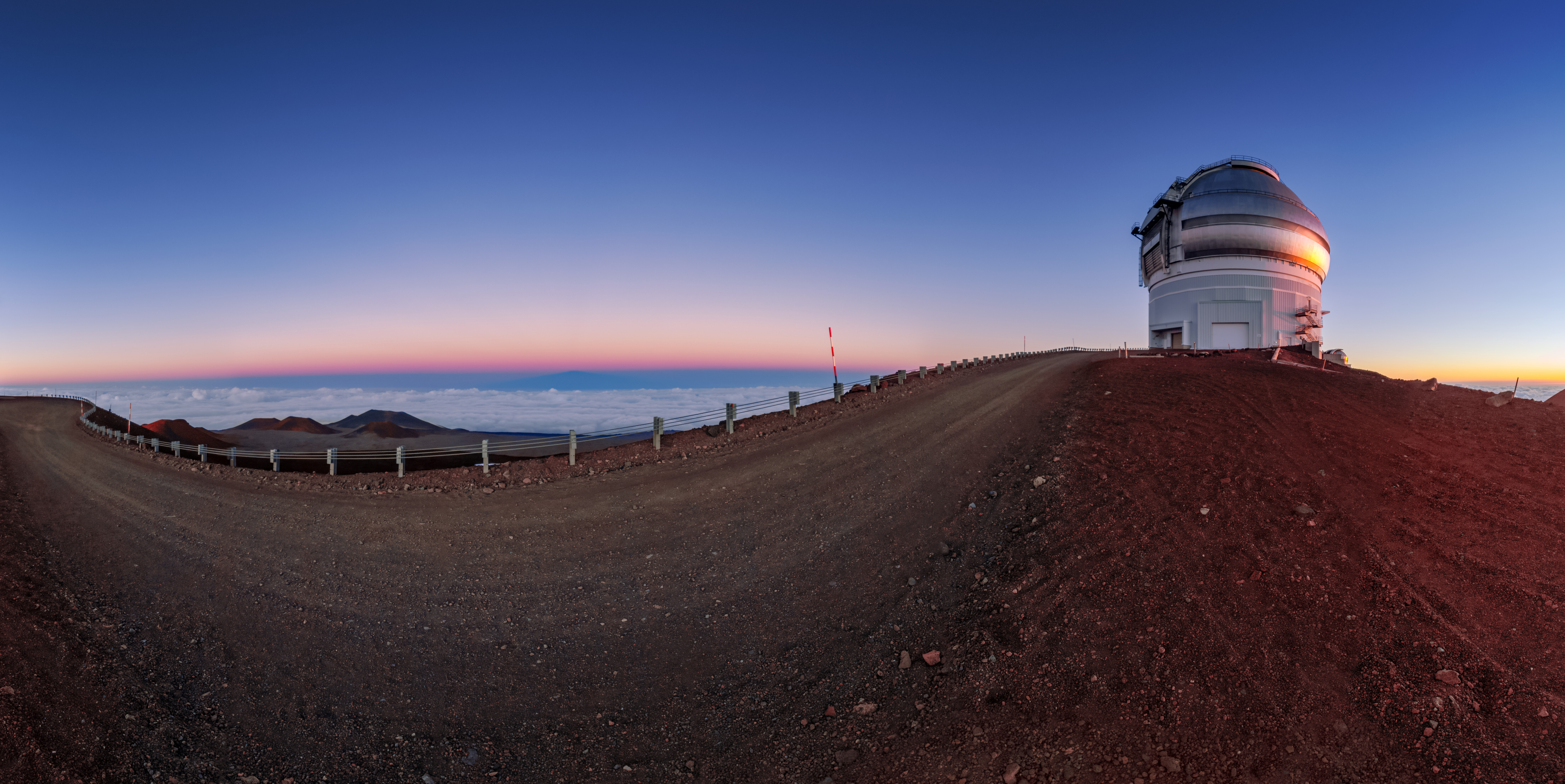 The silver dome of the Gemini North telescope, one half of the International Gemini Observatory, operated by NSF’s NOIRLab, catches the setting Sun, caught between the red-hued earth of the Maunakea volcano and an inky-blue sky. The scale of astronomical infrastructure can be difficult to grasp from images, particularly when telescopes are located in vast landscapes under wide skies. However, if you look closely the staircases winding up the side of Gemini North are visible — revealing the true size of this telescope. The dome of Gemini North — and that of its twin, Gemini South in Chile — is around 46 meters (150 feet) across, meaning that the two telescopes side-by-side would almost fit in an entire soccer field!This photo was taken as part of the recent NOIRLab 2022 Photo Expedition to all the NOIRLab sites.