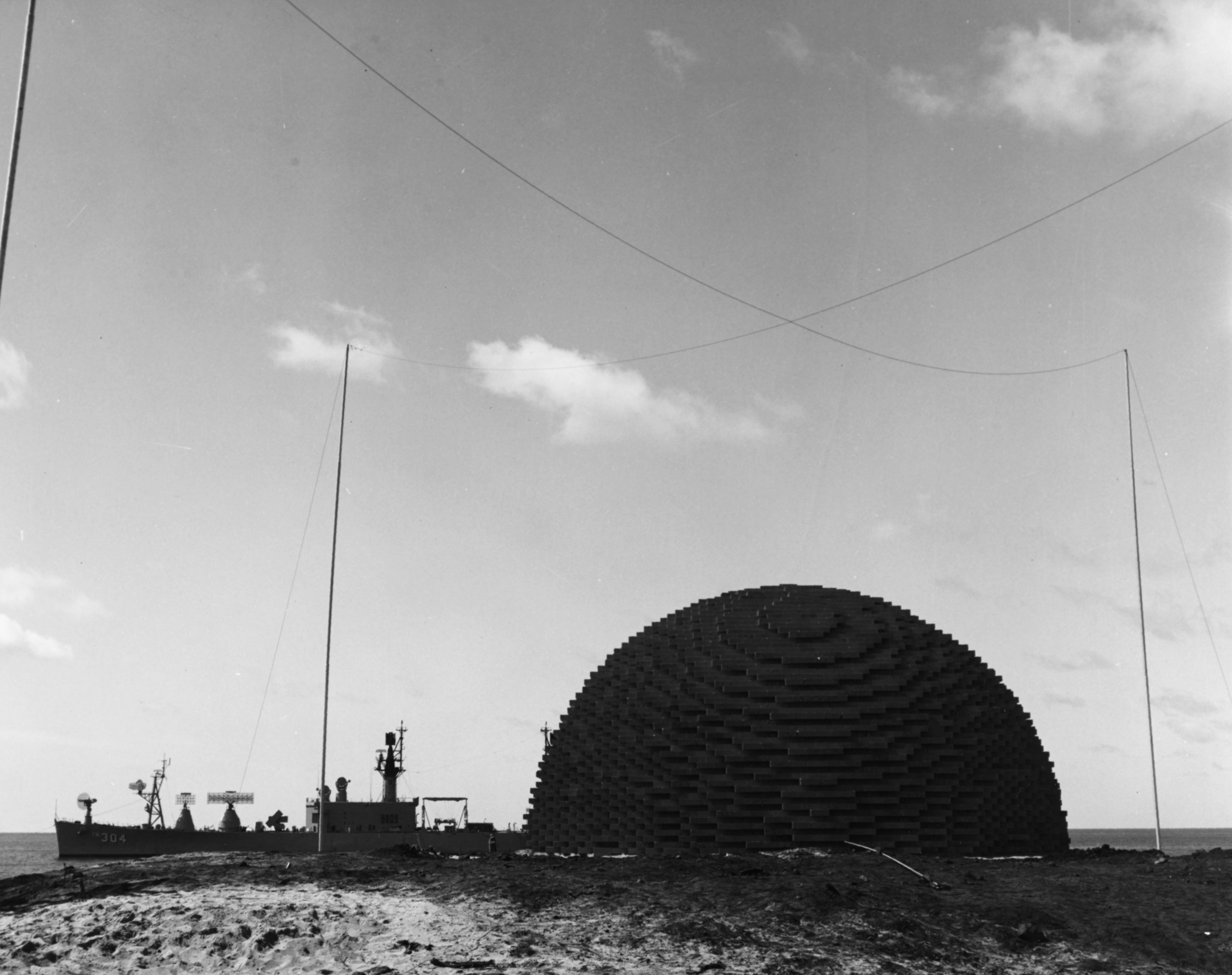 Operation Sailor Hat, 1965: The 500-ton TNT explosive charge for Shot Charlie, second of a series of three test explosions, ready for detonation on the southwestern tip of Kahoolawe Island, Hawaii, April 1965. USS Atlanta (IX-304) is moored in the background, with her bow facing left. A large SPS-37/SPS-43A type radar antenna is mounted on a pedestal on her foredeck, immediately in front of an ASROC launcher. An SPS-30 type radar antenna is on the pedestal at her extreme bow. Both of these antennas were relocated after the February 1965 Shot Bravo.