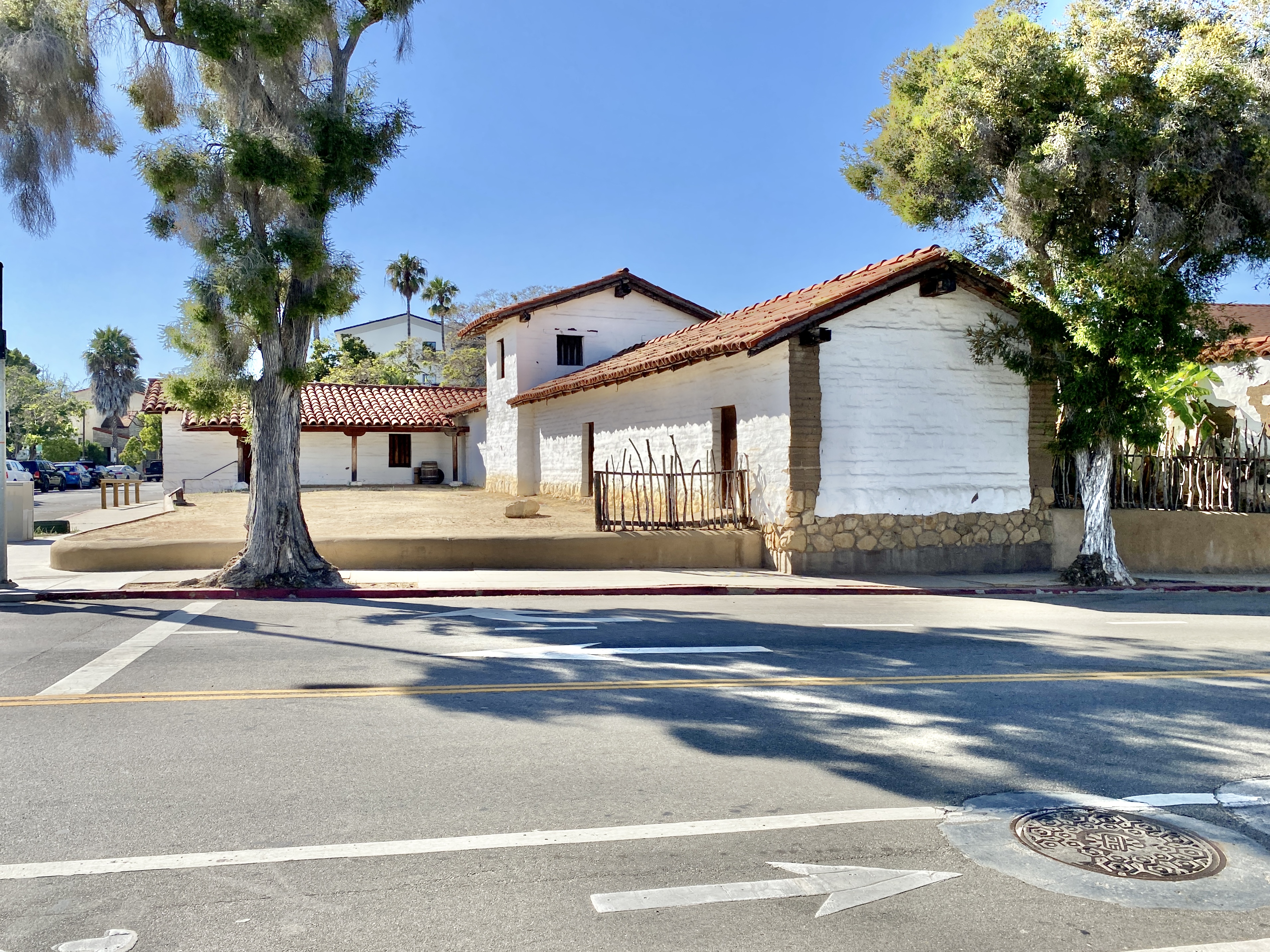 Originally built in 1782, this complex of buildings were constructed by the Spanish Colonial Authorities of California to serve as a presidio to protect the nearby Mission Santa Barbara and serve as a center of political influence in the surrounding region. The adobe buildings stood into the mid-19th Century, when they were slowly demolished and replaced by a neighborhood with roads platted through the middle of the presidio in the 1850s. By the mid-20th Century, only a single structure, the El Cuartel, which is the second-oldest existing building in California, remained standing, having been transformed into a private residence. The buildings were largely reconstructed after an archaeological investigation, with the first part of the complex being rebuilt between 1983 and 1985. The buildings consist of one-story adobe structures with red terra cotta tile roofs, exposed rafter tails, porches with wooden columns, and small window openings surrounding a central dirt courtyard, which has been partially restored, and a larger and taller chapel at the center of the western end of the courtyard, with a gabled roof, an arched entrance bay, and small tower with two bells. The buildings are divided by modern streets laid out after the Presidio had been largely demolished, with the ends of the reconstructed buildings revealing the interior structure of the presidio buildings where they are cut away along the streets. The original buildings in the complex were listed on the National Register of Historic Places in 1973. Today, the reconstructed Presidio serves as a historical museum operated by the Santa Barbara Trust for Historic Preservation.