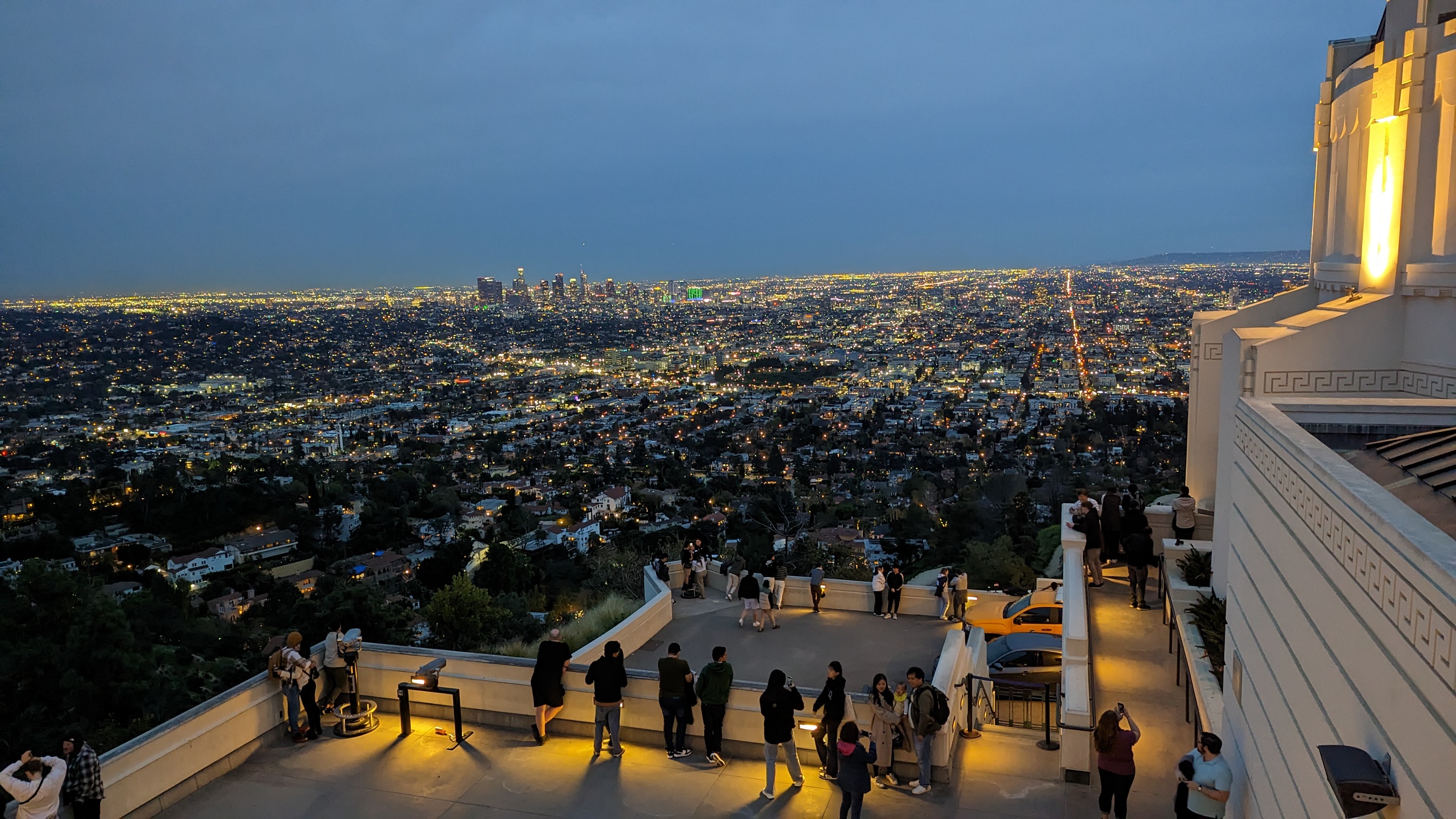Los Angeles as seen from the Griffith Observatory roof around dusk.  The lights of the city can be seen in the photo as well as the walls and buildings of the Griffith Observatory, along with several tourists taking in the sites on the rooftop below.  Taken in March of 2018 by Rachel L. Umstead during a site visit to the Getty Center.