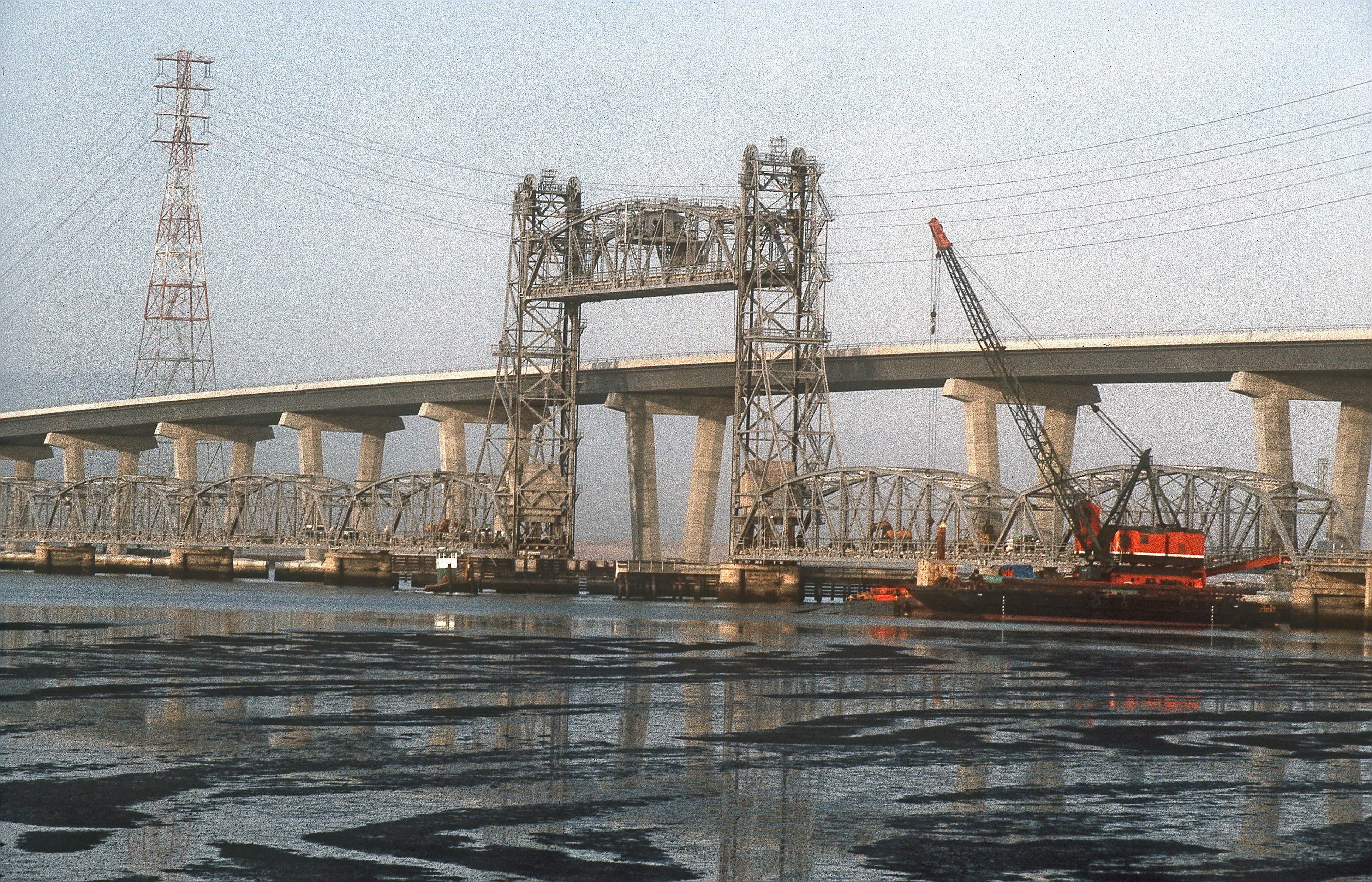 The original vertical-lift Dumbarton Bridge is shown alongside its replacement span, shortly before the demolition of the older bridge in September 1984. The bridge is the southernmost crossing of the San Francisco Bay.