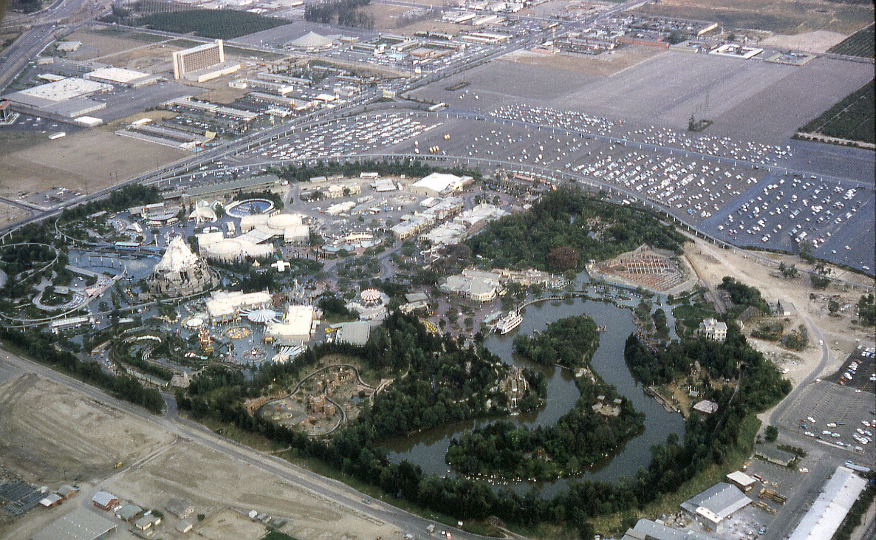 Aerial view of Disneyland looking southeast in 1963, located in Anaheim, California, United States. The Santa Ana Freeway (I-5) is in the upper left corner. Harbor Boulevard forms the eastern boundary of the park. To the middle right, one can see the Haunted Mansion under construction. While the main building is complete on the outside (the white building), the show building is just beginning construction on the right side of the railroad tracks (most of the ride takes place in a show building outside the confines of the railroad). The Haunted Mansion would open in 1969. Also under construction, and up and left from the Haunted Mansion, is Pirates of the Caribbean, which opened in 1967. The two attractions took a while to build because Disney's engineering arm at the time, WED Enterprises (now known as Walt Disney Imagineering), was busy on projects dealing with the 1964 New York World's Fair. Both attractions are located in New Orleans Square, which opened in 1966. Anaheim's newly completed Melodyland Theater ("theater-in-the-round"), is at the top of the photo.