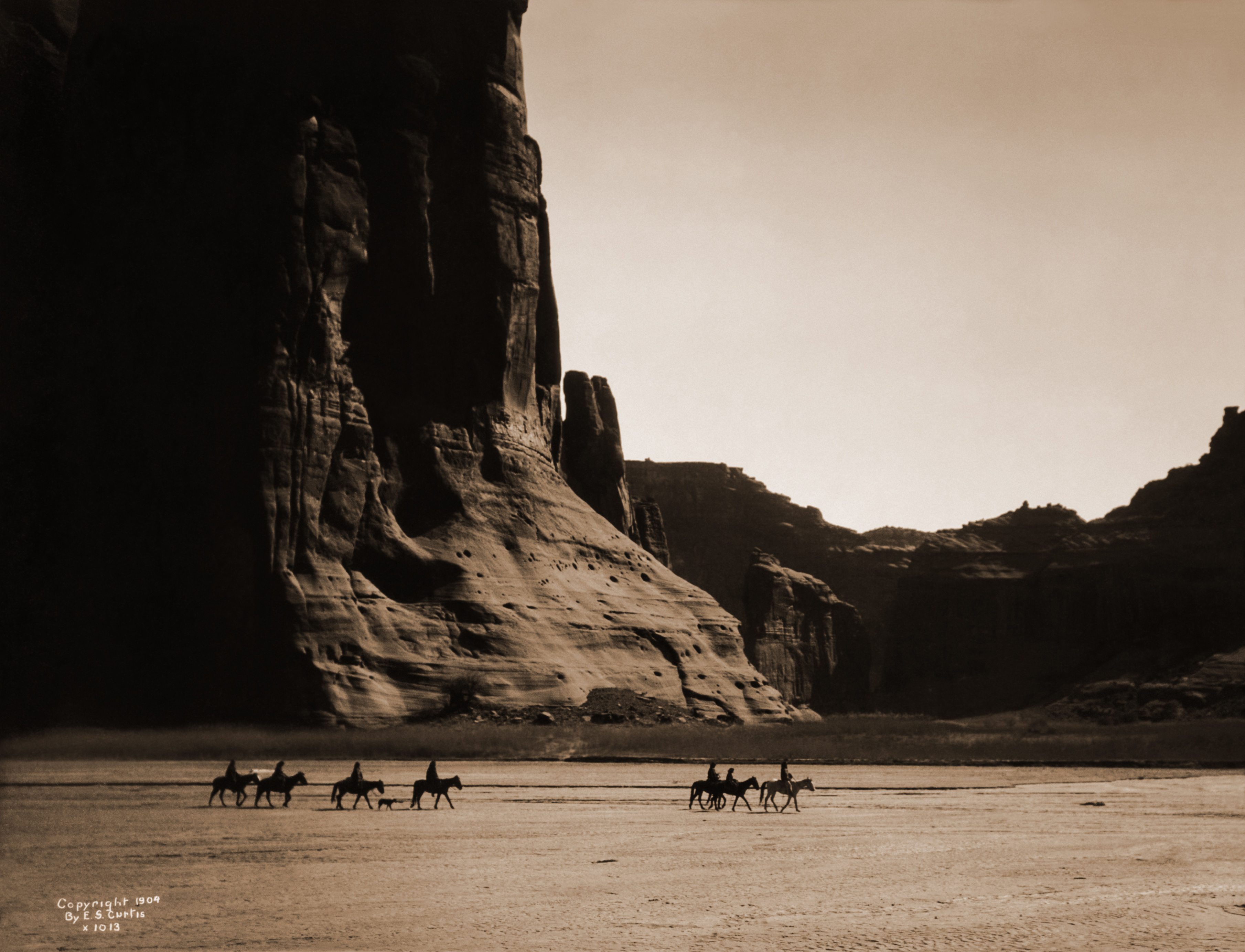 E. S. Curtis (1904): Canyon de Chelly – Navajo. Seven riders on horseback and dog trek against background of canyon cliffs.