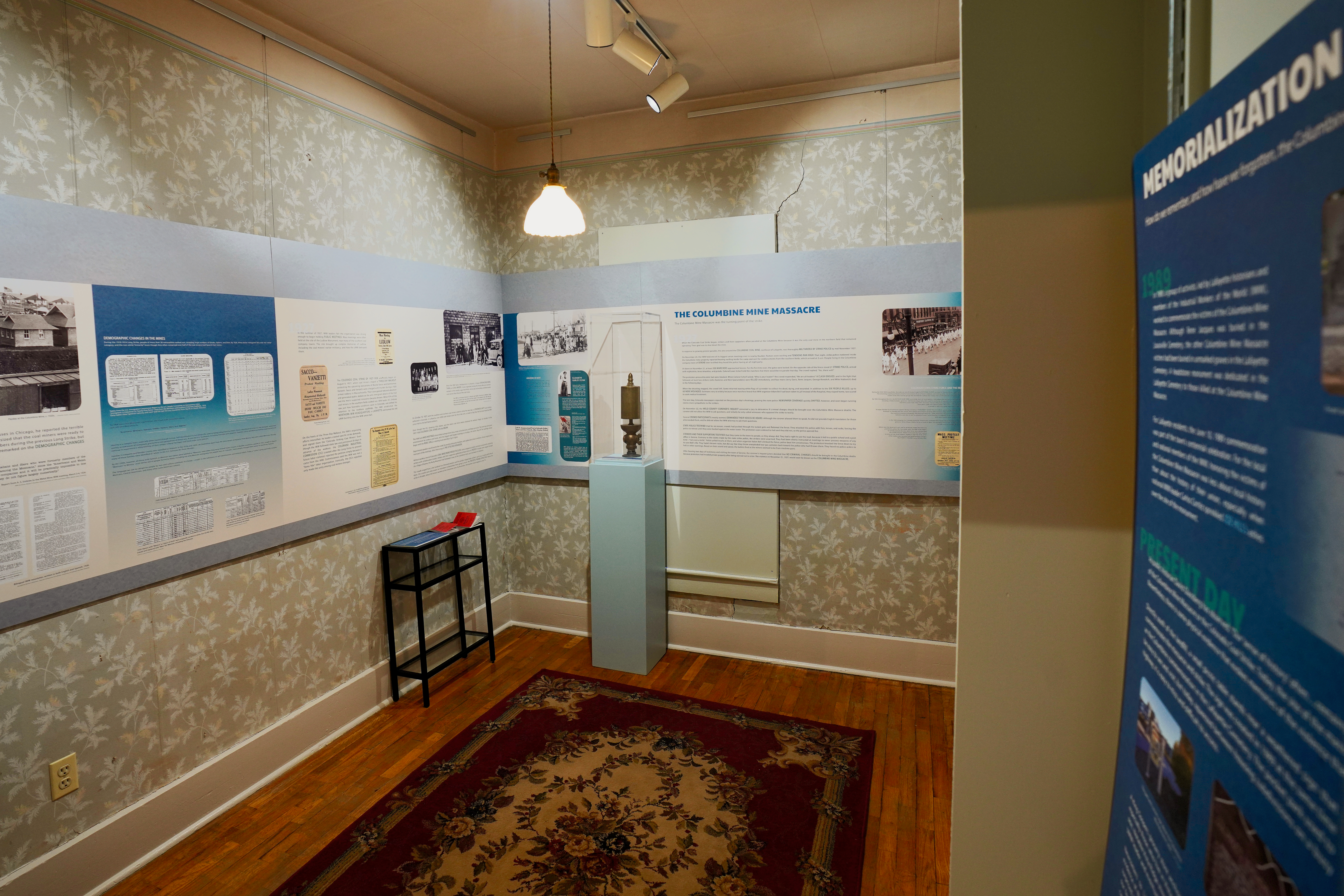 Photograph of a Lafayette History Museum exhibit on the Columbine Mine Massacre, showing interpretive panels, historical photographs, documents, and a mining lamp on display. Taken inside the museum in Lafayette, Colorado.