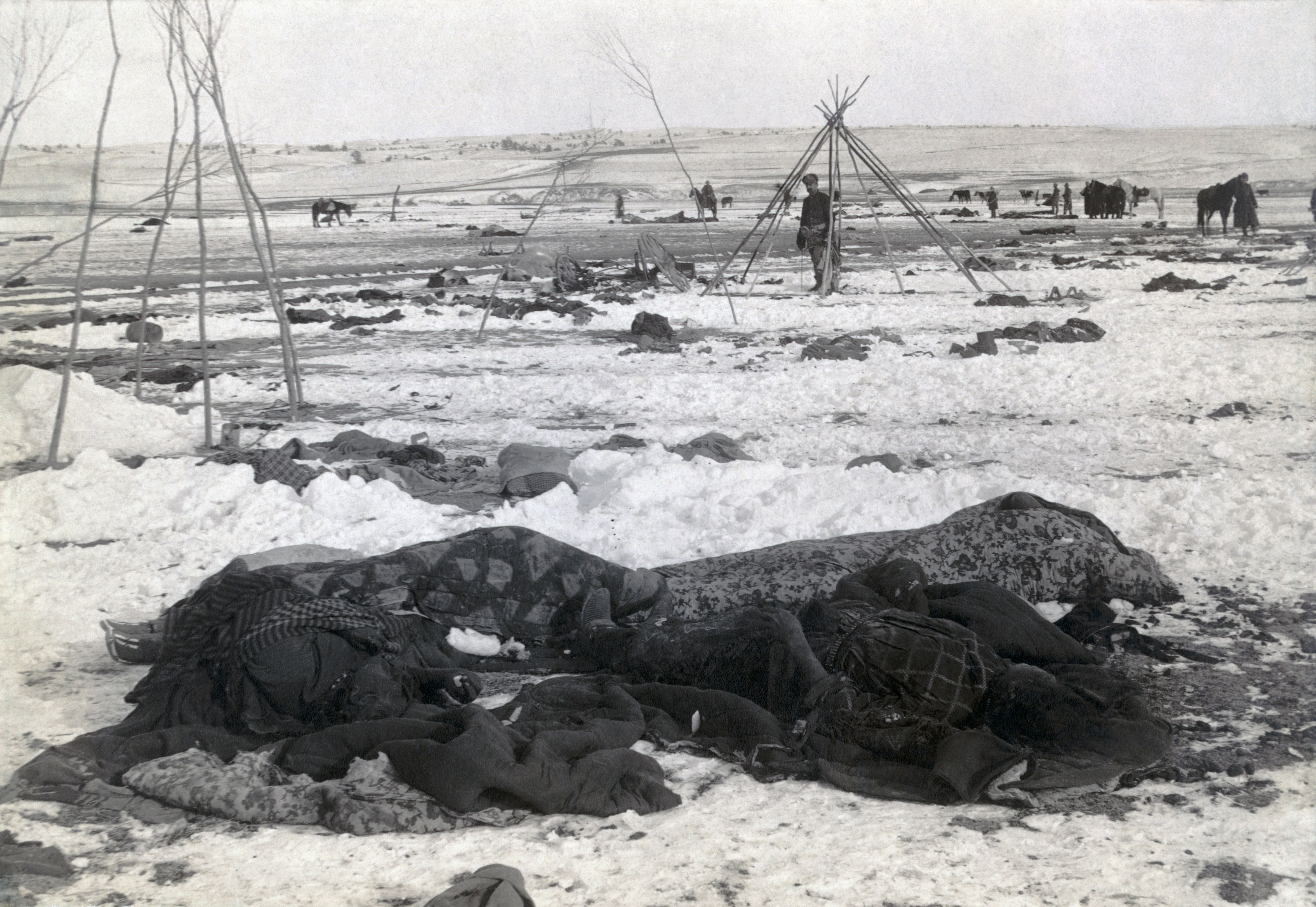 Bibliographic caption: "Big Foot's camp three weeks after Wounded Knee Massacre; with bodies of four Lakota Sioux wrapped in blankets in the foreground; U.S. soldiers amid scattered debris of camp"   1 photographic print : albumen ; 9 x 11 in.