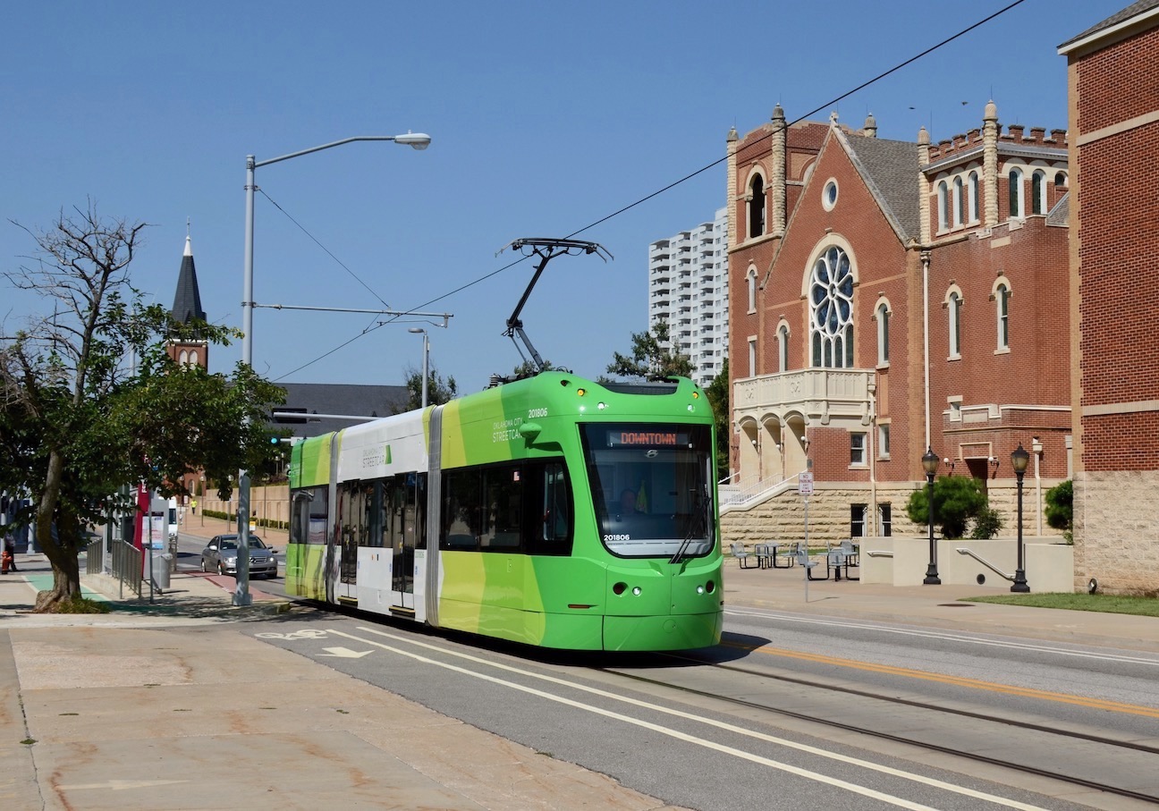 Green-and-white car No. 201806 of the Oklahoma City Streetcar system eastbound on NW 4th Street just east of Robinson Avenue, passing the First United Methodist Church. Car 201806 was built by Brookville Equipment Corporation for the OKC Streetcar system, which opened in 2018. The historic church building was built in 1903 as the First Episcopal Methodist Church. It was damaged in the 1995 bombing of the nearby Murrah Federal Building and was subsequently restored.