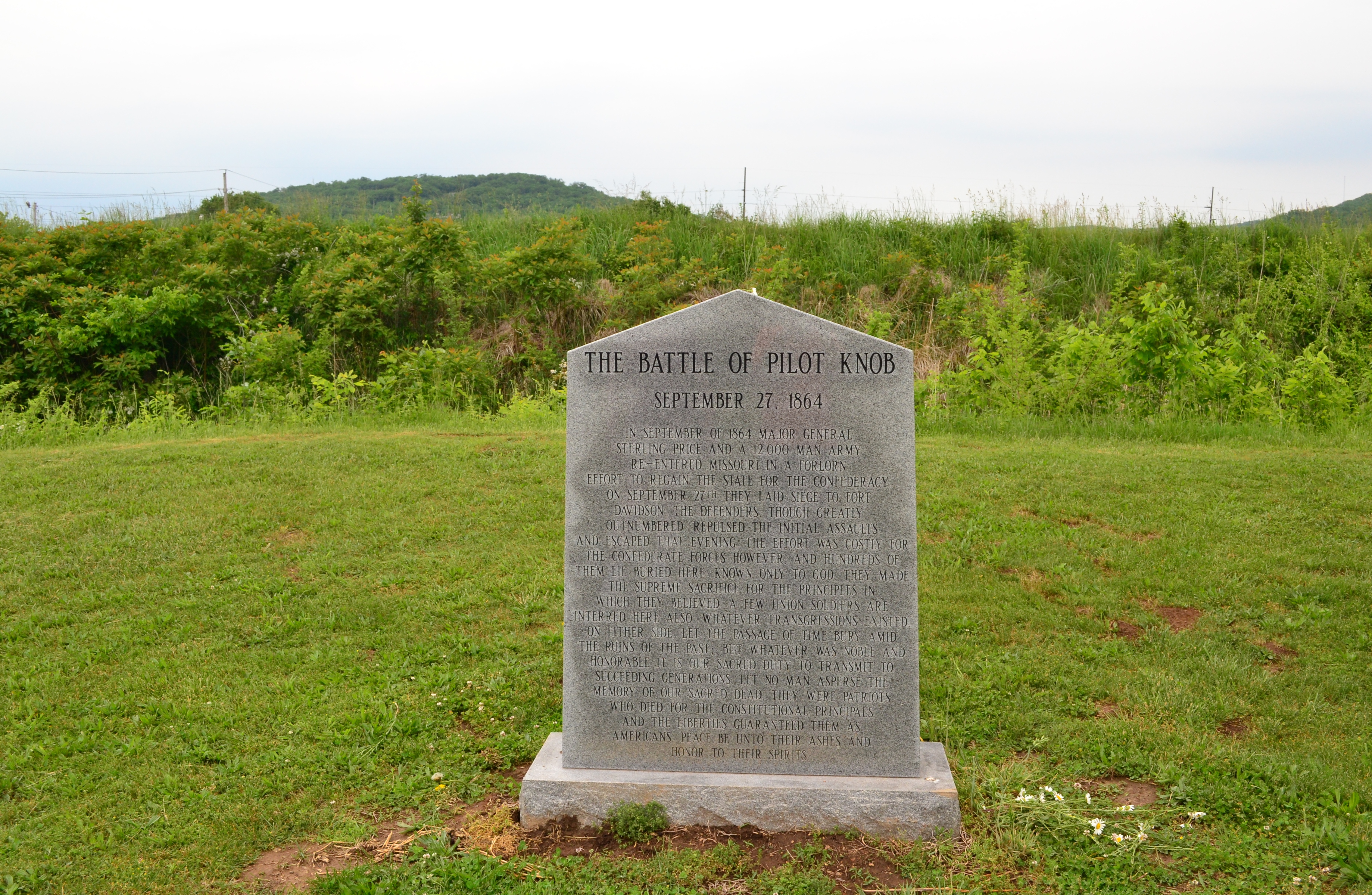 A stone memorial monument at Fort Davidson in Missouri.  The earthen rampart of the fort's ruins is behind the monument.
