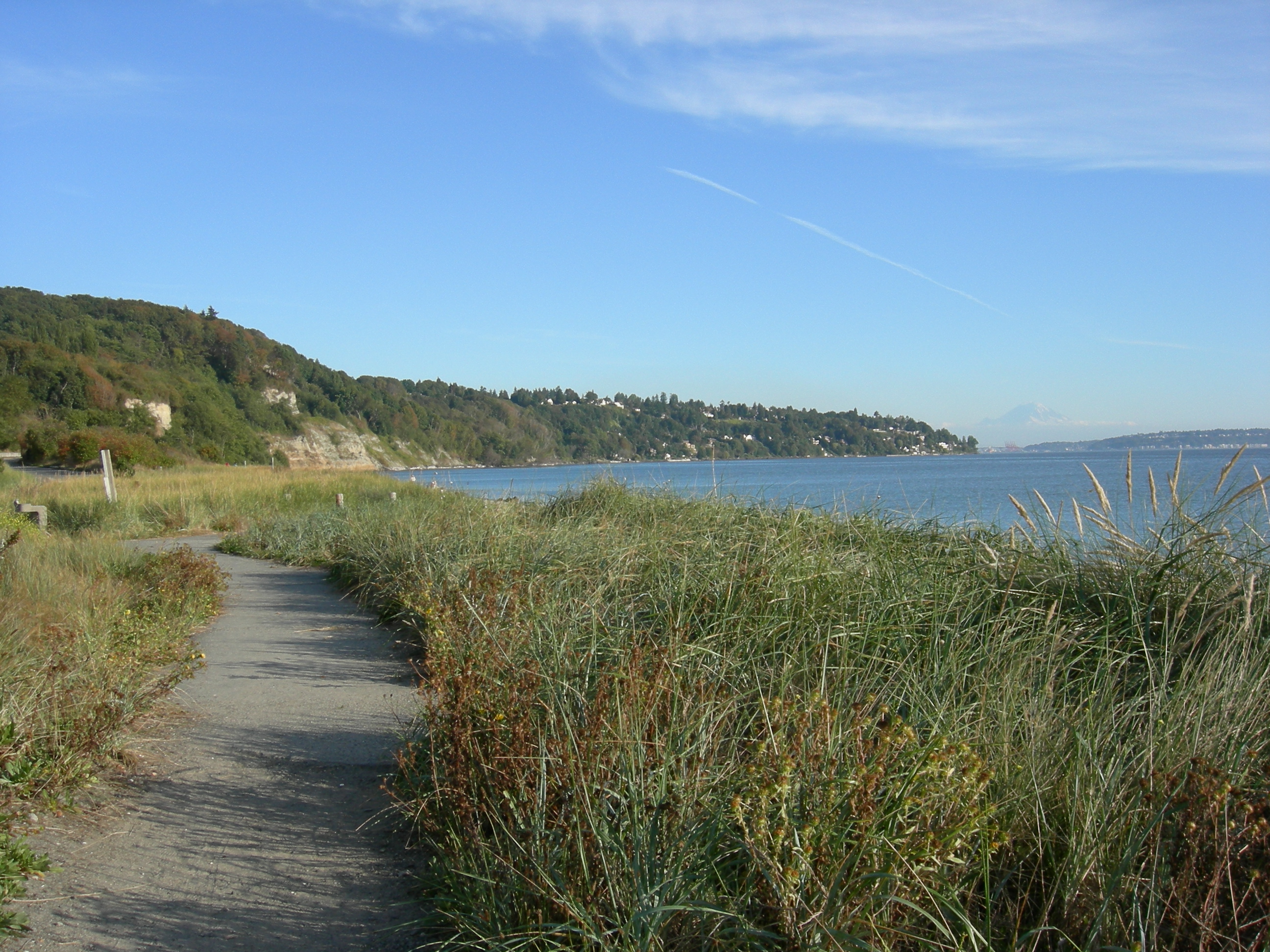 Looking roughly southeast from West Point along the path along the south beach of Discovery Park, Seattle, Washington, USA. Beyond the beach are sand cliffs of the park itself, then (to the right) houses further south on Magnolia. In the distance at left are Mount Rainier and West Seattle. The body of water is the northwest corner of Elliott Bay, part of Puget Sound.