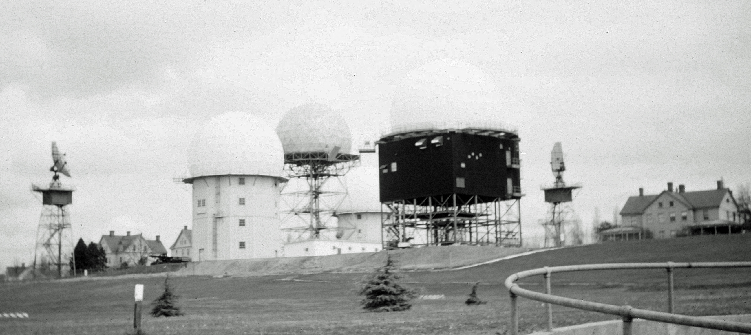 A multi-agency radar site located at Fort Lawton, Washington, circa 1962-63. The radome in the center with the uncovered steel structure is a search radar owned and maintained by the Federal Aviation Administration. The two radar systems on the outside of the photo, without radomes, are US Army FPS-6 height-finders. The two radars with radomes and white covered structure beneath (one near, the other in the back) are US Air Force FPS-6A height-finders. The radar with a white radome and dark covered structure beneath is a US Air Force FPS-26A height-finder, under construction in this photo.