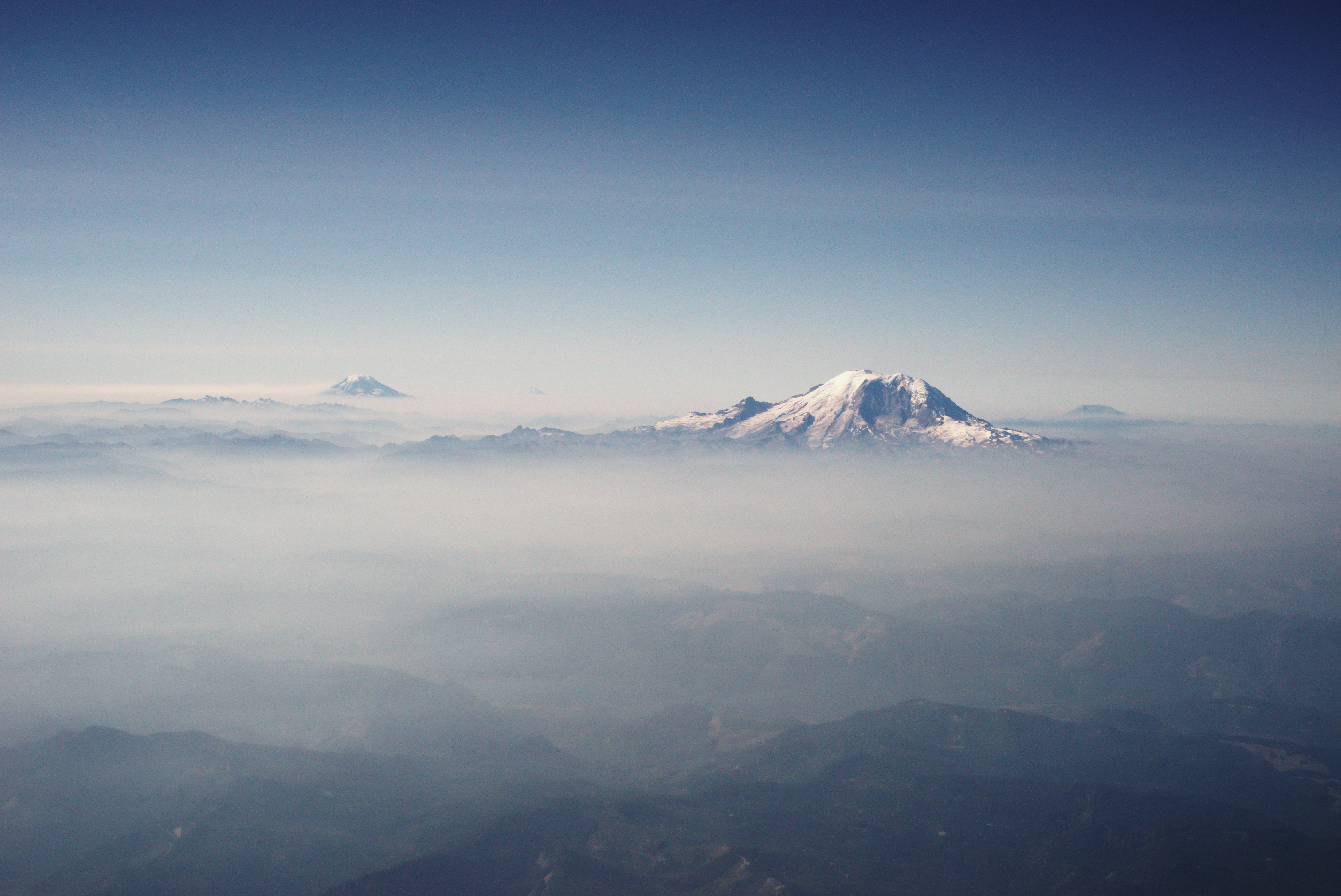 Four Cascade Range mountains (left to right): Mount Adams, Mount Hood (in Oregon), Mount Rainier (foreground), and Mount St. Helens, seen poking through the cloud layer shortly after takeoff on a commercial flight from Seattle to Detroit. Despite bringing a microfiber cloth to clean the aircraft window, the poor optical quality of the window resulted in a blurry photo.