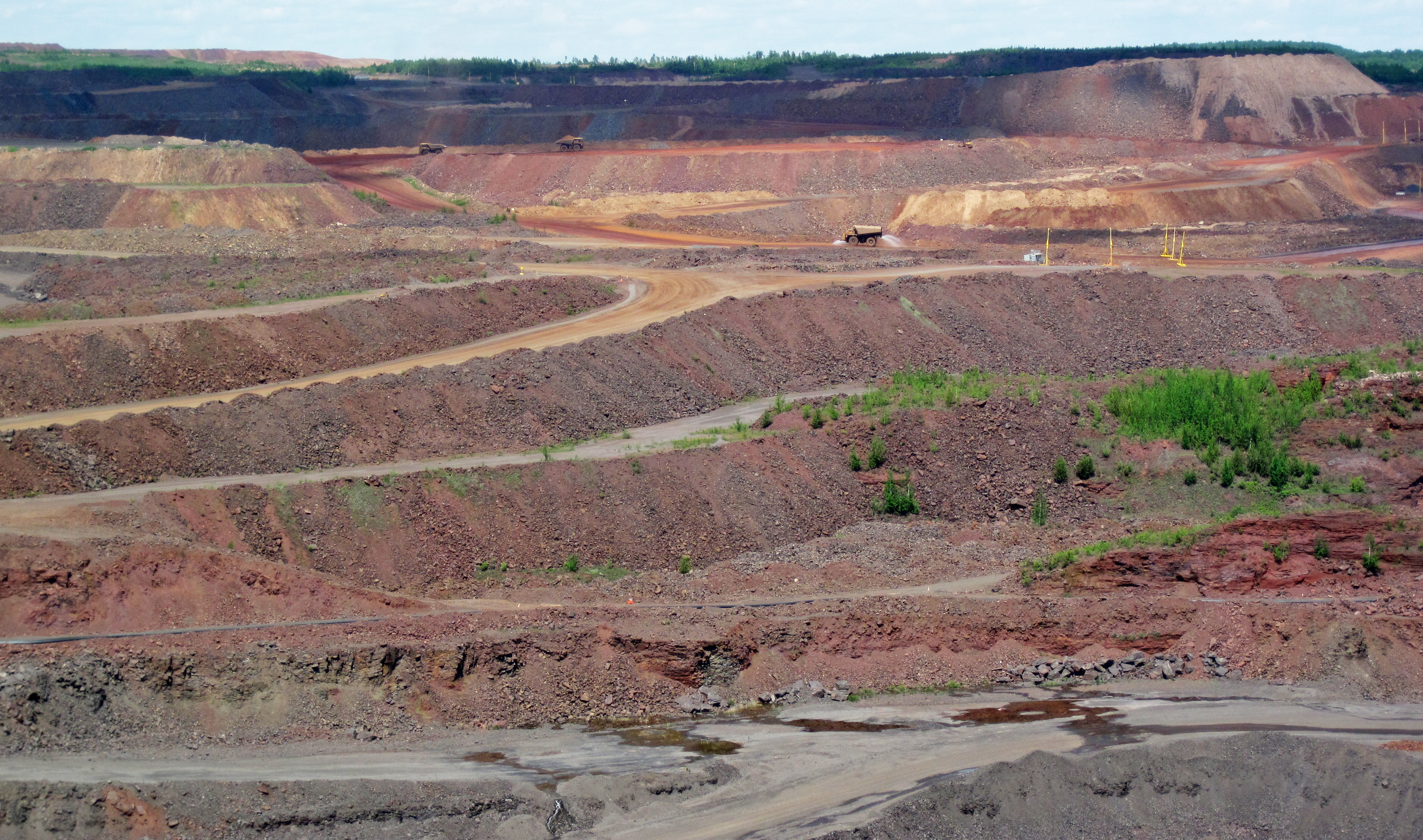 Banded iron formations, or BIFs, are unusual, dense, marine sedimentary rocks consisting of alternating layers of iron-rich oxides and iron-rich silicates.  Most BIFs are Proterozoic in age, although some are Late Archean.  They do not form in today's oceans - they're an example of an “extinct” rock type.  Many specific varieties of iron formation are known.  The most attractive type is jaspilite, which is a reddish &amp; silvery gray banded rock consisting of hematite, red chert (“jasper”), and specular hematite or magnetite.  Most BIFs have been subjected to one or more orogenic (mountain-building) events, resulting in folding and/or metamorphism.  BIFs are known from around the world, but some of the most famous &amp; extensive deposits are found in the vicinity of North America’s Lake Superior Basin.  Many BIFs have economic concentrations of iron and are mined as iron ores.
The Hull-Rust-Mahoning Mine shown above is an active iron mine in Minnesota's Mesabi Iron Range.  Multiple mines have operated at this site and have since merged into one giant operation, which is currently a pit ~5 miles long and ~1.5 miles wide.  Mining started in the mid-1890s and continues today.  A significant percentage of America's iron needs during World War I and World War II were met by production from this single mine.
The target rocks at the Hull-Rust-Mahoning Mine are taconites in the Precambrian-aged Biwabik Iron-Formation, which is the iron ore producing unit throughout the Mesabi Iron Range.  Early mining operations focused on the weathered zone of the Biwabik Iron-Formation, which is rich in earthy hematite and low in silicates.  Modern mining produces taconite as iron ore, which is relatively rich in silicates but still has significant hematite content.
Stratigraphy: Biwabik Iron-Formation, Paleoproterozoic, ~1.878 Ga

Locality: Hull-Rust-Mahoning Mine, northern side of the town of Hibbing, Mesabi Iron Range, western St. Louis County, northeastern Minnesota, USA (47° 27' 16.59" North latitude, 92° 56' 42.87" West longitude)