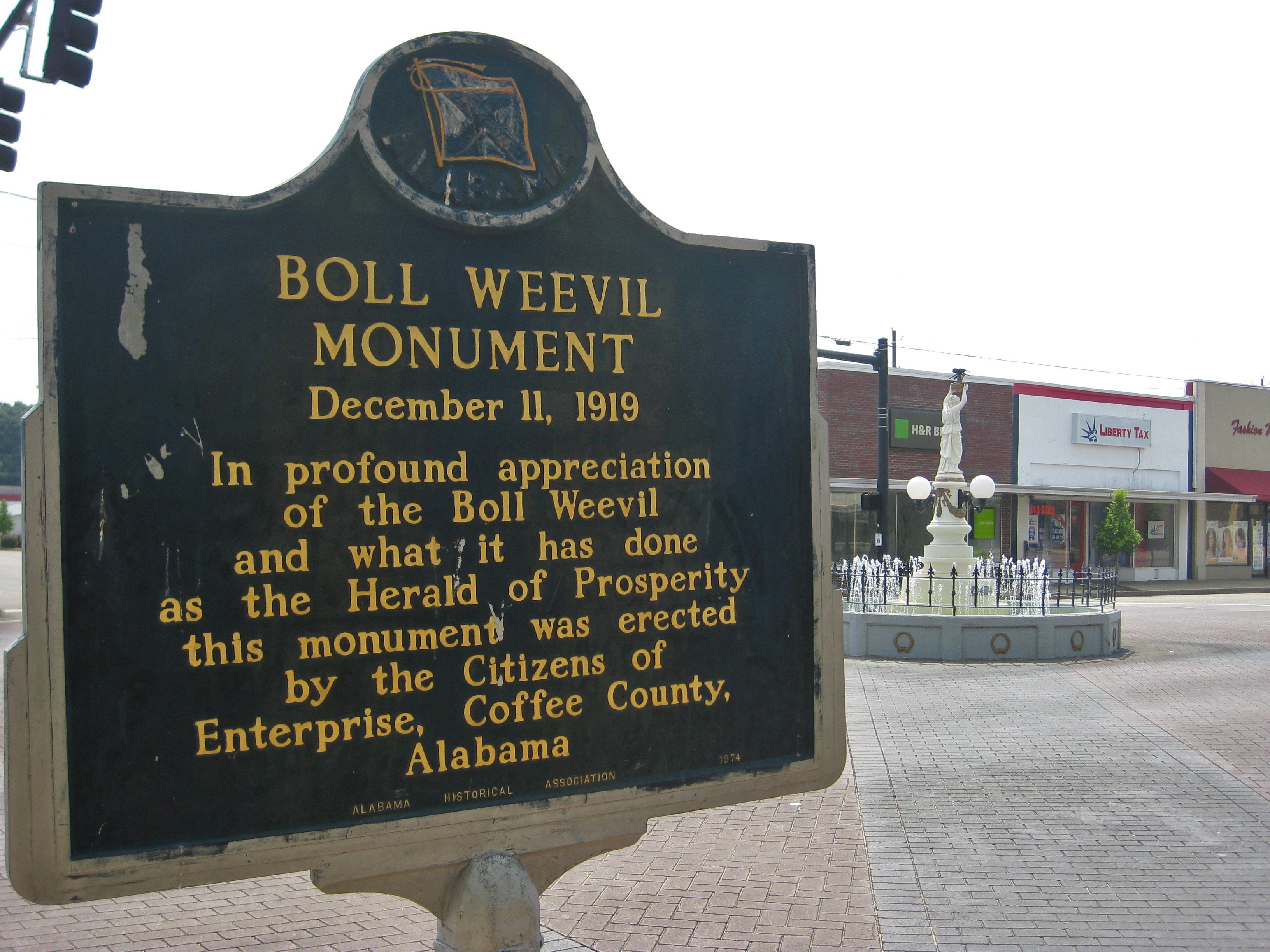Alabama Historical Association marker for the Boll Weevil Monument in Enterprise, with the Boll Weevil Monument in the background to the right.