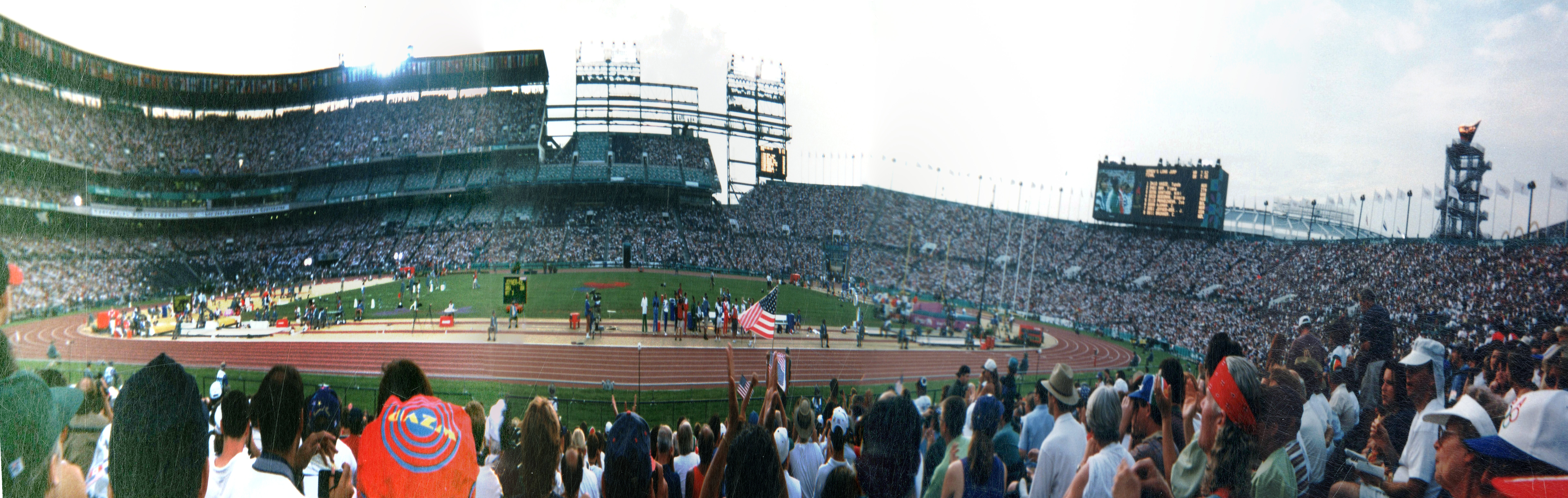 This is a panorama of Centennial Olympic Stadium during track and field events one hot summer afternoon near the end of the events, as seen from the east side of the stadium.  Jackie Joyner-Kersee was competing long jump that afternoon as evidenced by her name on the scoreboard near the track after her third attempt, and the other competitors are named on the large scoreboard to the right.
The panorama has been made by stitching together 4 photos that were developed, printed, scanned, and manually aligned, then stitched together and projected in Hugin Panorama Creator, hence the track doesn't always align.  A small triangle of sky was missing from the original and has been substituted with nearby data.