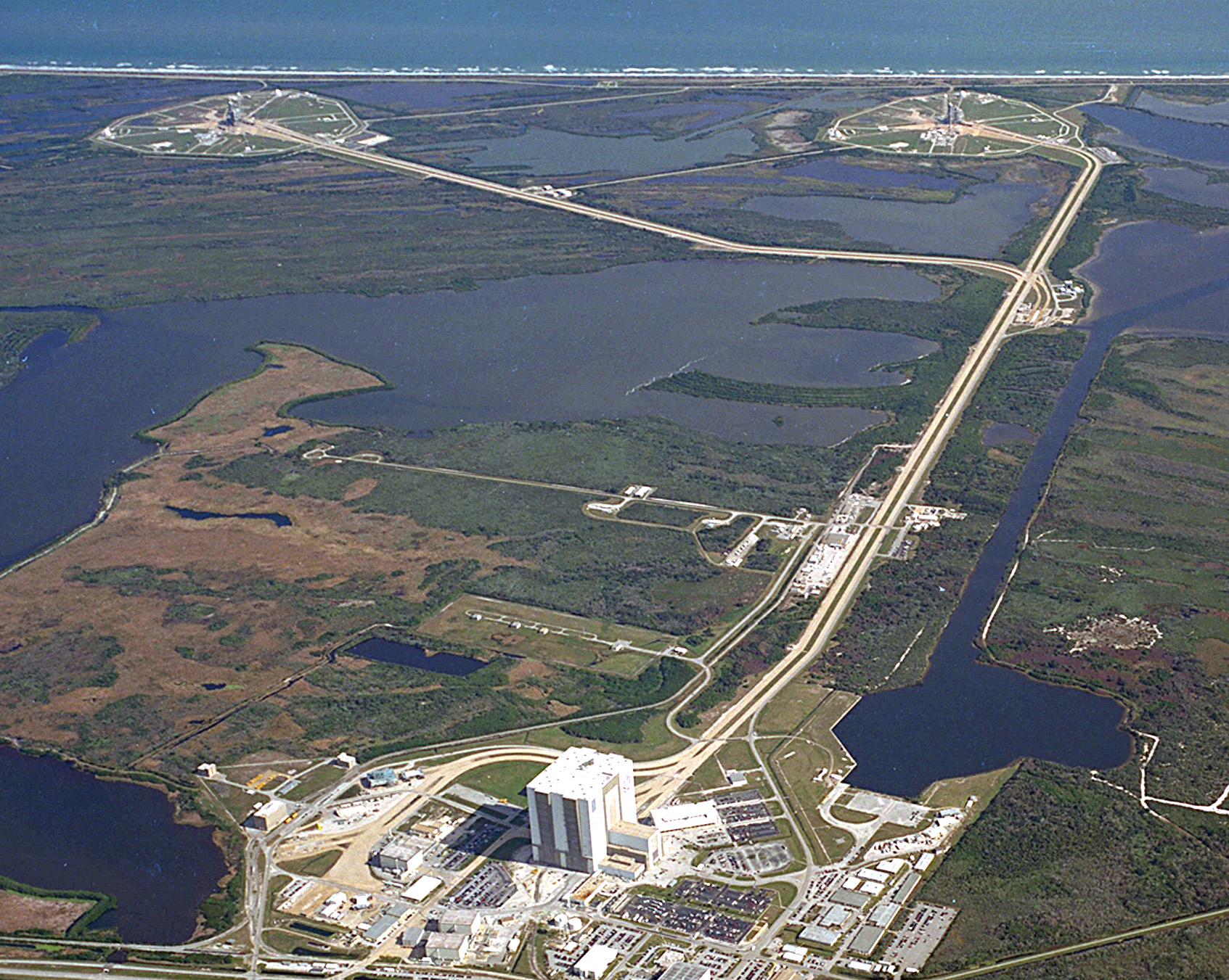 KSC's Launch Complex 39 is strategically located next to a barge site and a variety of structures, including a Vehicle Assembly Building (VAB), Orbiter Processing Facilities (OPF), Press Site, Launch Control Center (LCC), and a crawlerway to the pads.
The crawlerway, leading to pads 39B on the left and 39A on the right, can be seen extending from the massive VAB at the left in this photo. The VAB, which covers eight acres and stands 525 feet tall, is used for assembly, stacking and mating of Space Shuttle elements. Originally built for assembly of Apollo/Saturn vehicles and later modified to support Space Shuttle operations, the VAB is one of the largest buildings in the world.
The LCC, seen here as the small white building to the upper right of the VAB, is where launch, mission support, and loading are controlled.