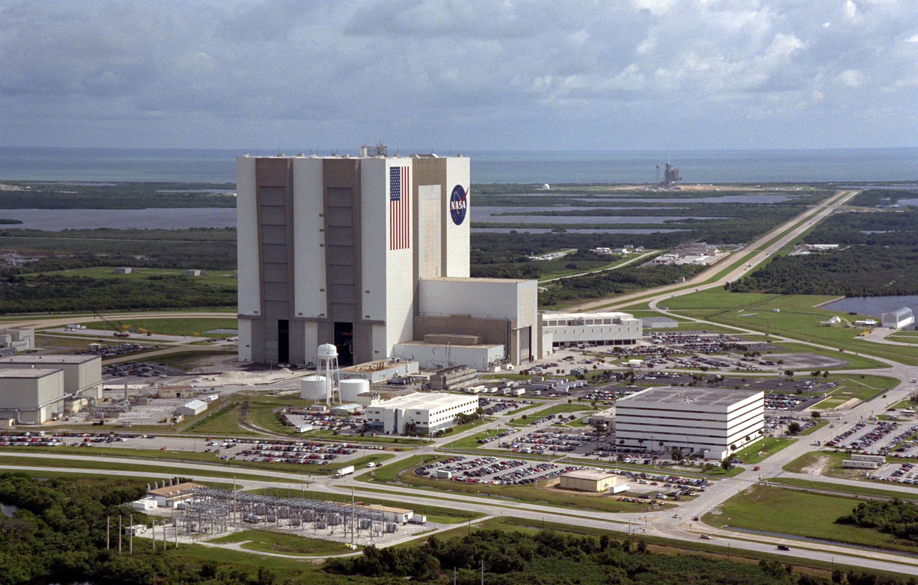 Kennedy Space Center - Aerial View of Launch Complex 39An aerial view of the Launch Complex 39 area shows the Vehicle Assembly Building (center), with the Launch Control Center on its right. On the west side (lower end) are (left to right) the Orbiter Processing Facility, Process Control Center and Operations Support Building. Looking east (upper end) are Launch Pads 39A (right) and 39B (just above the VAB). The crawlerway stretches between the VAB and the launch pads toward the Atlantic Ocean, seen beyond them. At right is the turning basin where new external tanks are brought via ship.