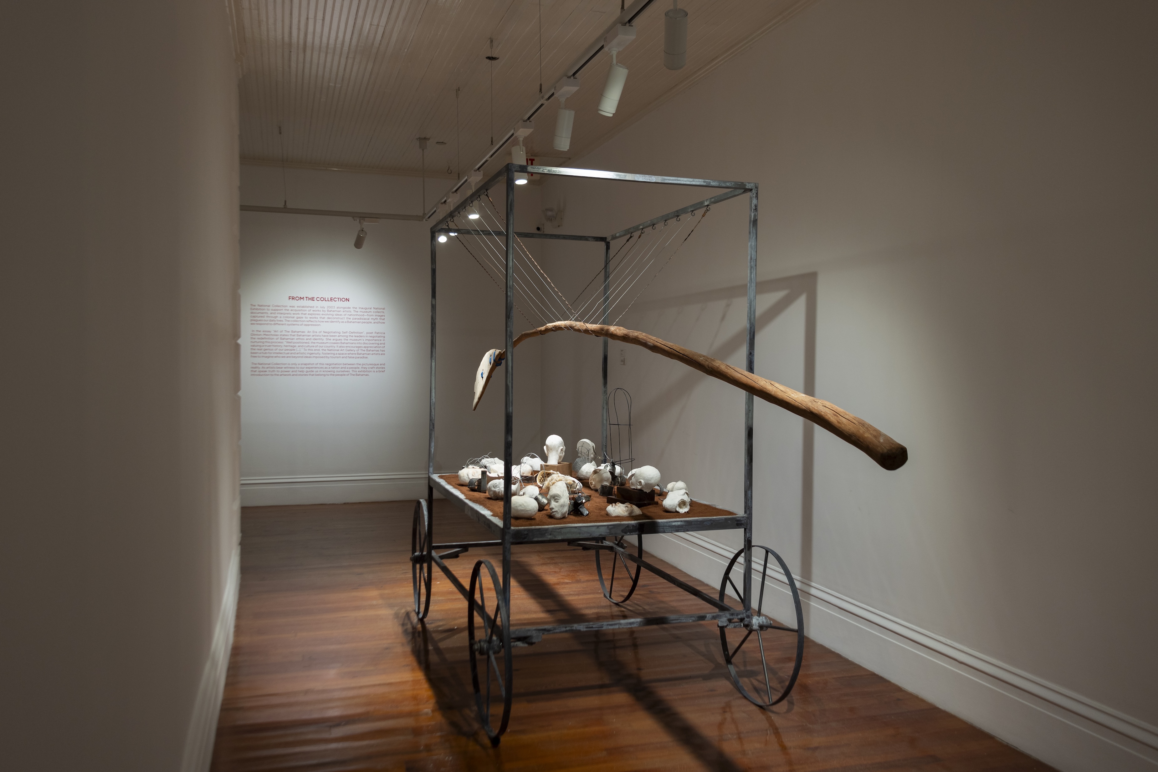 A mixed media installation by John Beadle featuring an iron frame with wheels that supports a wooden oar, several skulls, and a bed of red sand. John Beadle was born in 1964 on the island of New Providence in the Bahamas