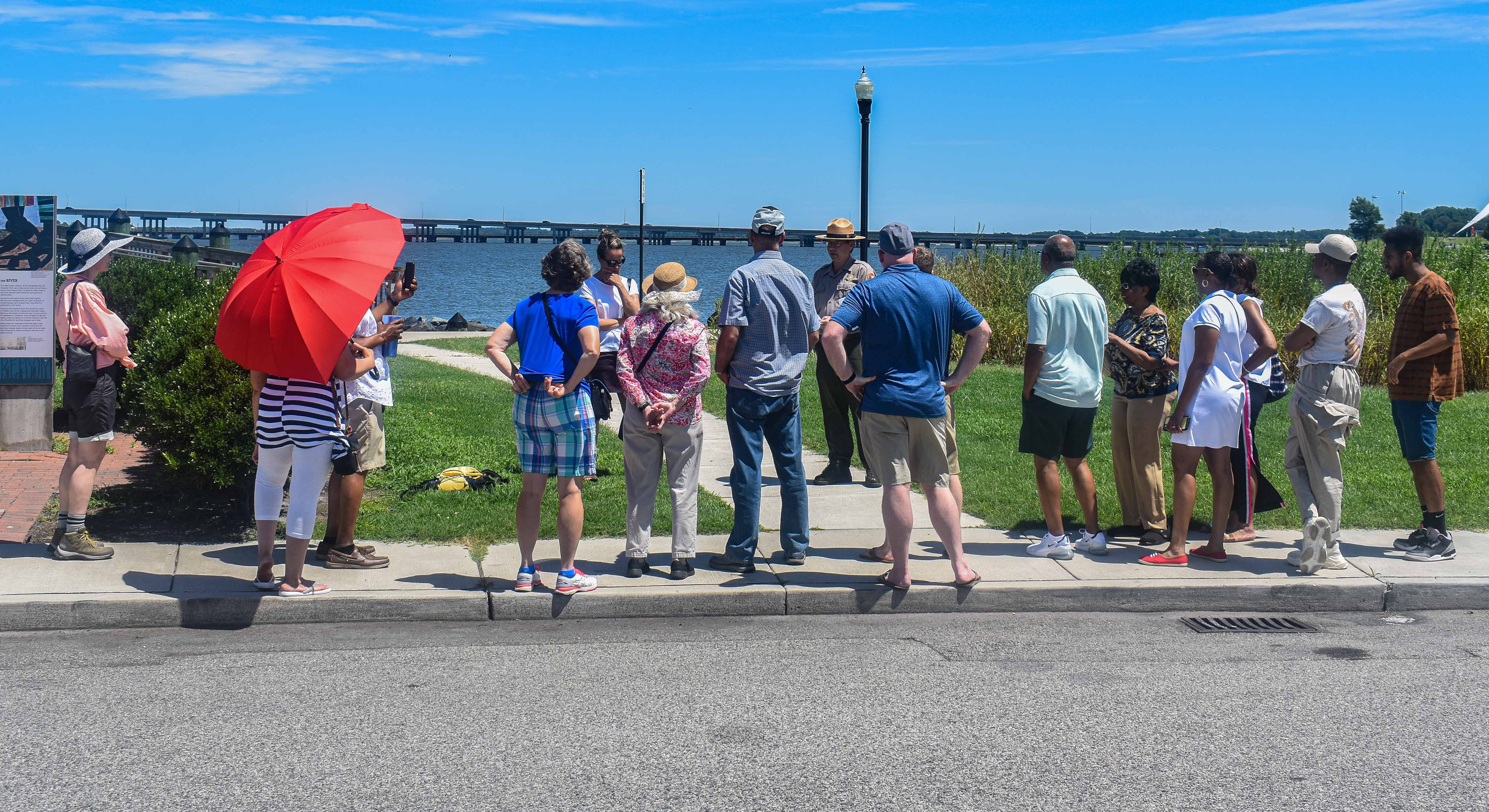 In the foreground, an asphalt road can be seen. An NPS Ranger can be seen presenting a piece of paper while giving an interpretive talk to a group of 13 guests standing on a sidewalk at historic Long Wharf in Cambridge, MD.  A light post, the Choptank River, and the Choptank Bridge can be seen in the background.
An NPS ranger gives an interpretive talk at historic Long Wharf in Cambridge, MD.
Keywords: Harriet Tubman; Harriet Tubman Underground Railroad National Historical Park; ranger program; walk; photography