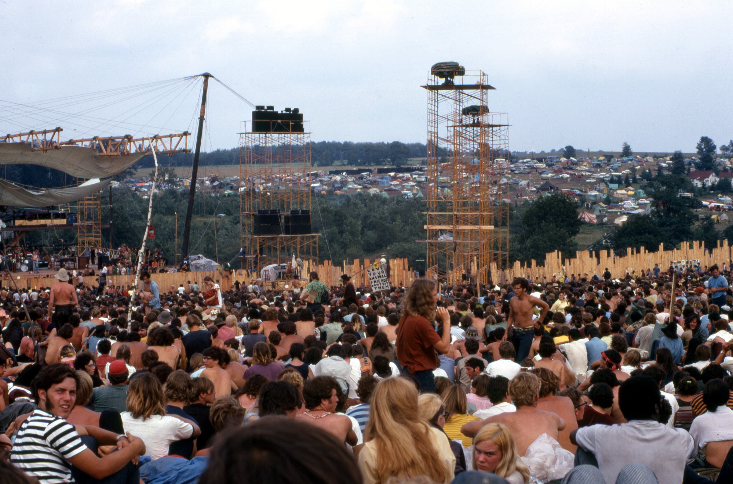 Picture taken on 17 August 1969 at the Woodstock Music and Art Fair. The person carrying the placard is Moonfire Lewis Beach Marvin III. Joe Cocker is performing on stage.