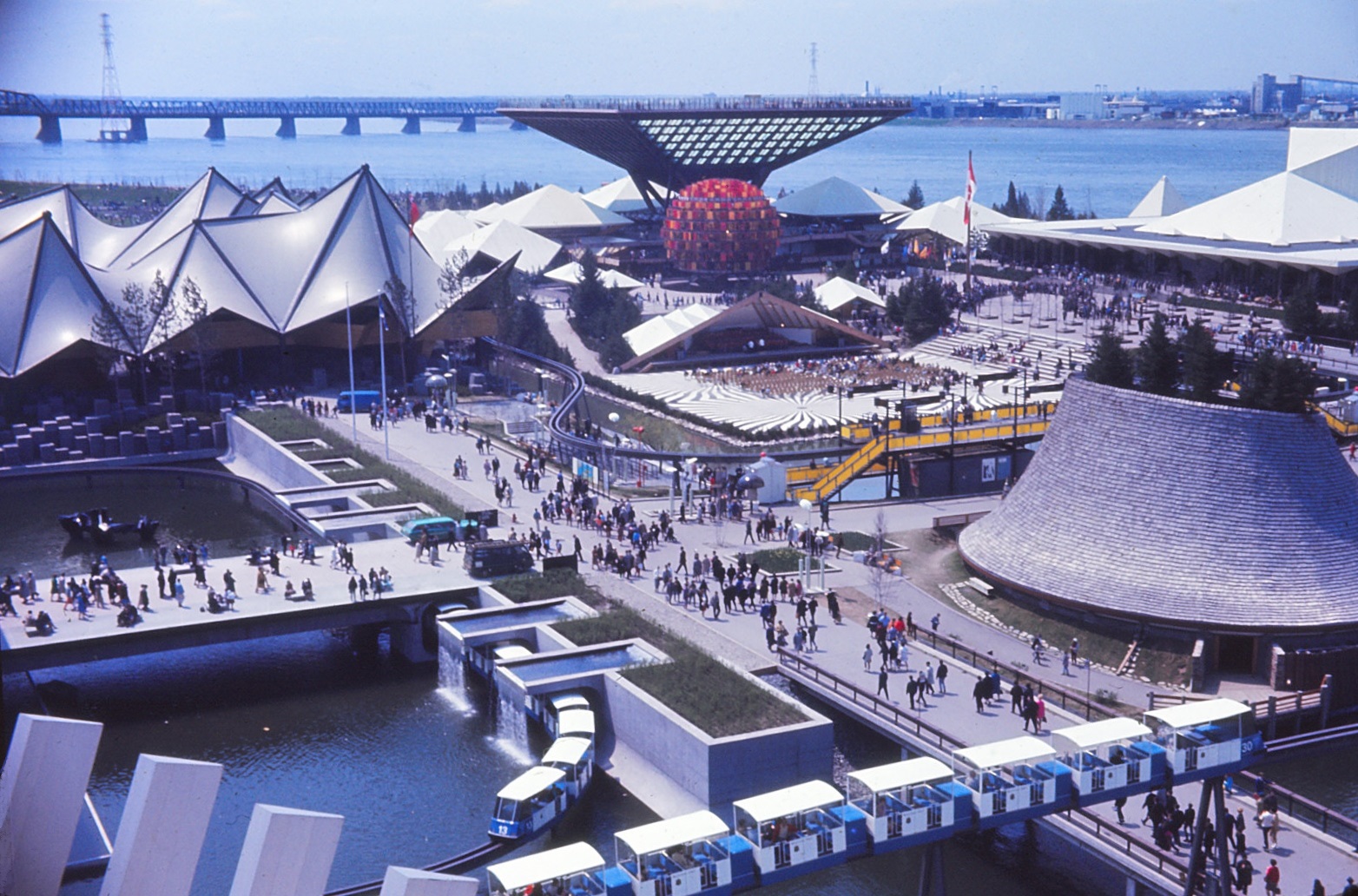 Pavilions of Ontario, Canada, Western Provinces, seen from the pavilion of France. On the left, the basin and waterfalls of the Quebec pavilion. One can see the Minirail surrounding the pavilions. Expo 67, Montreal, Quebec, Canada.