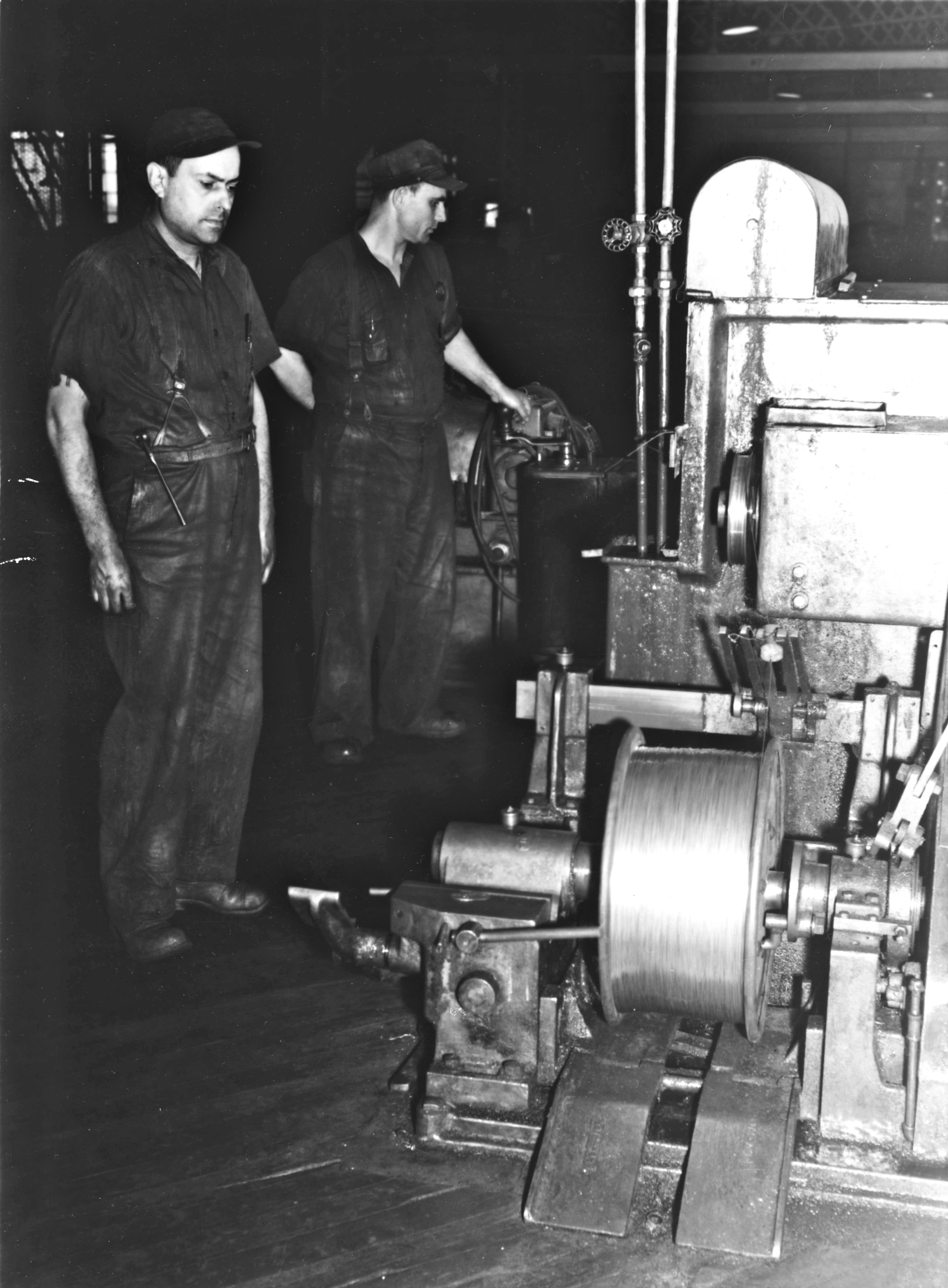 Travailleurs de la tréfilerie, usine 1, Alcan, Shawinigan. Une version un peu rognée de cette photo a été publiée dans La Revue de l'aluminium, Shawinigan Falls, vol. III, no 6, édition du 17 mars 1945, en page 6, où la légende de la photo précise que les deux tréfileurs sur cette photo sont Armand Arsenault et Donat Houle, qui surveillent la réduction du diamètre du fil d'aluminium, qui entrera ensuite dans la fabrication de câbles d'aluminium. L'article associé mentionne que les câbles d'aluminium sont produits pour les lignes de transmission électrique.