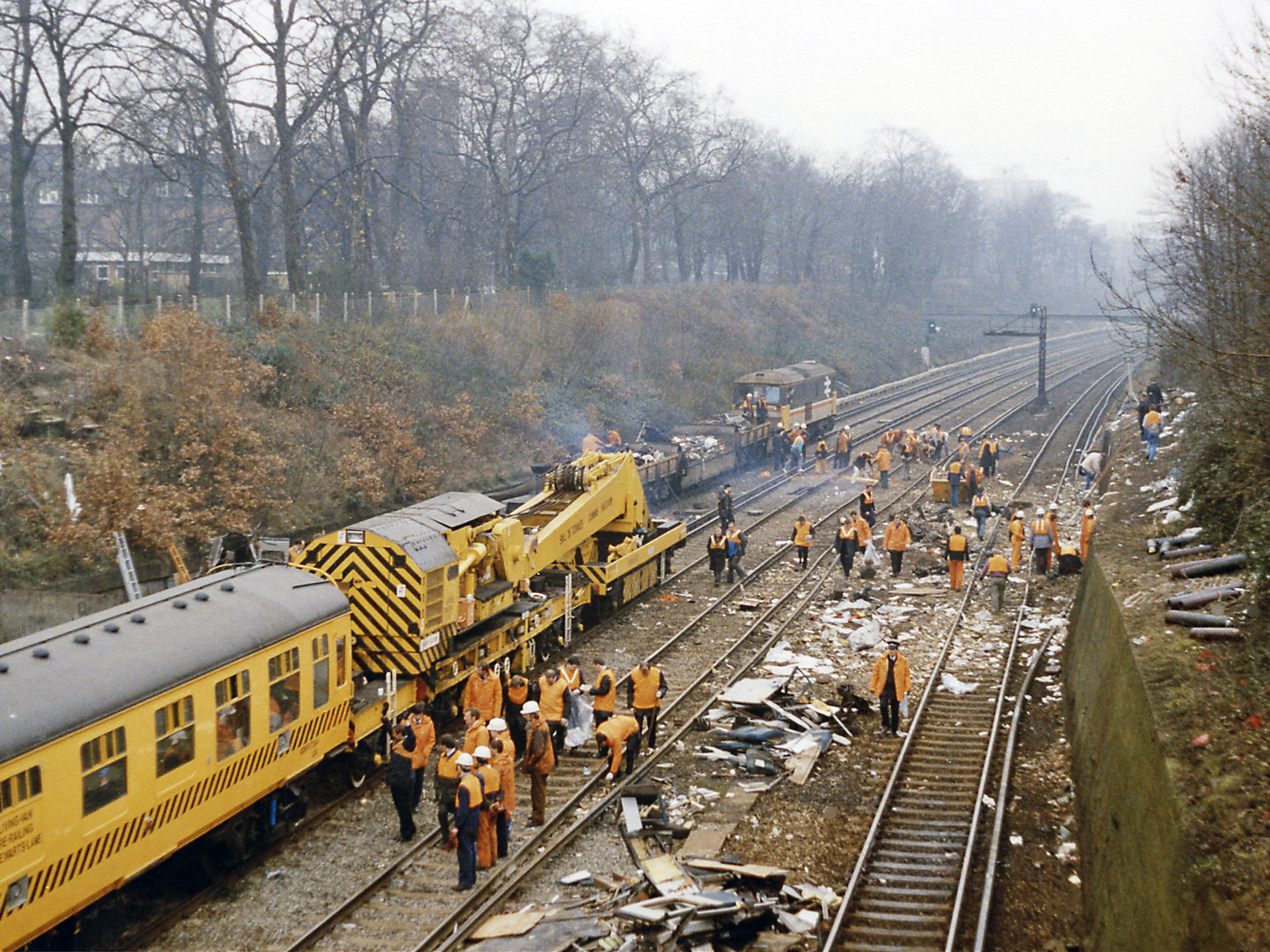Clearing up after the December 1988 crash west of Clapham Junction.
View SW towards Wimbledon etc. from the Battersea Rise bridge/Spencer Park Road on the day after a bad accident had occurred on the Up lines (nearest) at 08.10 the previous morning, in which 35 people were killed and over 500 injured. Owing to an installation fault with the signalling, a crowded Up train had run into the rear of another and the wreckage was then struck by a Down empty train. The view shows all four ex-LSWR tracks of the main Waterloo - Wimbledon - Woking and the West line blocked by the breakdown-train men at work more than 24 hours afterwards. The numerous casualties had been brought up the bank on the right to Spencer Park Road. (See also TQ2674 : Clearing up after the December 1988 crash west of Clapham Junction).