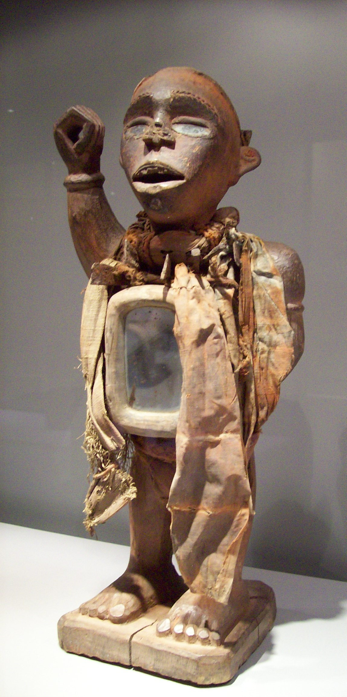 Magische Figur (Bakongo, Mayombe, Dem. Republik Kongo)
Die Figur weist sowohl ein Spiegelkästechen (nur an der Brustseite!) als auch einige eingeschlagene Nägel und Klingen auf. Das Gesicht ist fein ausgearbeitet, ein Fischgerätemuster betont die plastisch erhöhten Augenbrauen, die Lippen des leicht geöffneten Mundes sind sorgfältig geformt; die obere Zahnreihe zeigt die für die Yombe charakteristische Lückenaussplitterung. Die Kopfbedeckung (mit Zauberbehälter) fehlt.