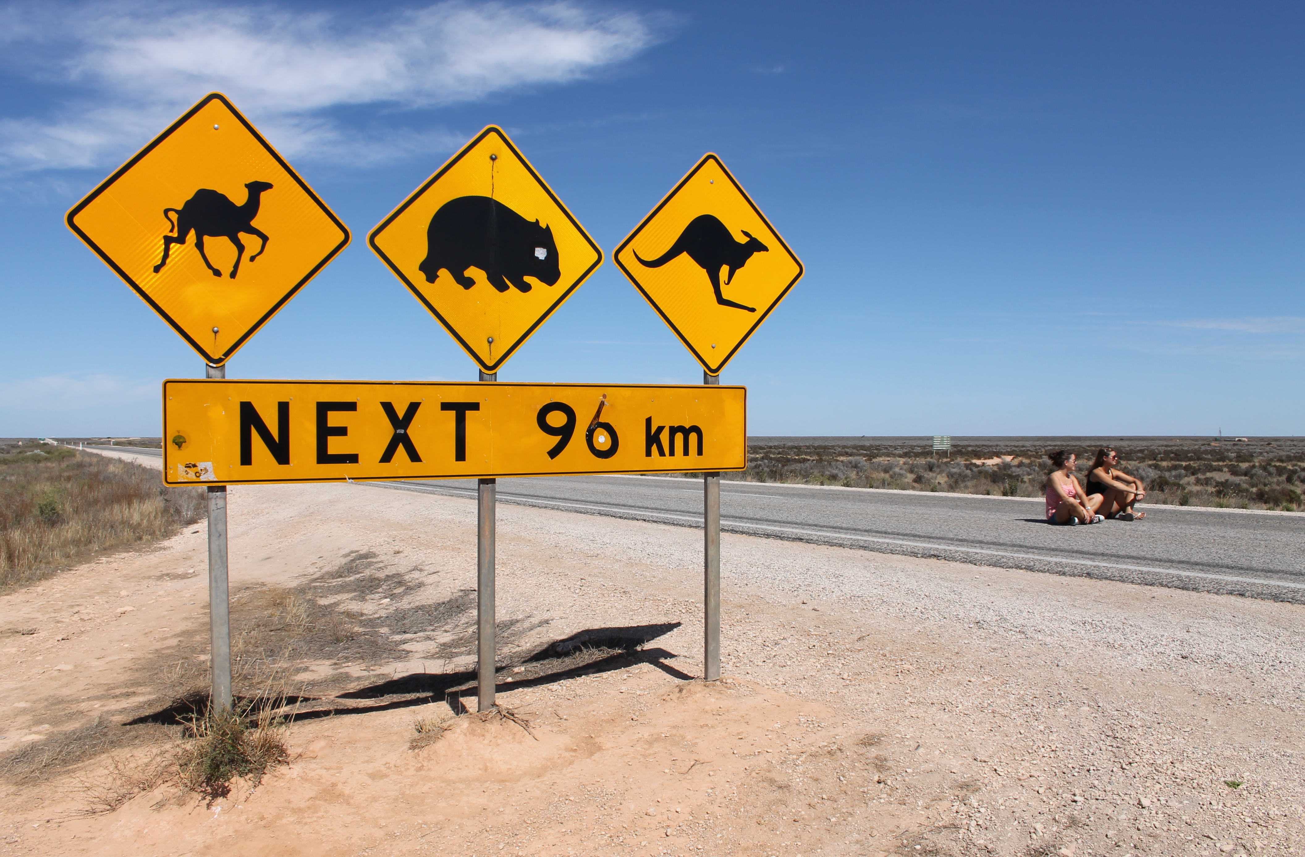 A set of road signs just west of the Nullarbor Roadhouse, South Australia, warning of camels, wombats and kangaroos crossing the Eyre Highway for the next 96 km in a westerly direction.  A couple of visitors to Australia, Hannah and Toni, are sitting on the highway at right.