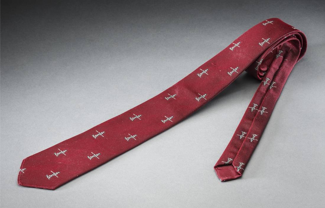 Maroon polyester necktie. It has a repeated motif of an Aboriginal man carrying a spear and a shield in silver and black bevo weave. 'Austico / TERYLENE / ICI' is on the label attached to the back of the tie.