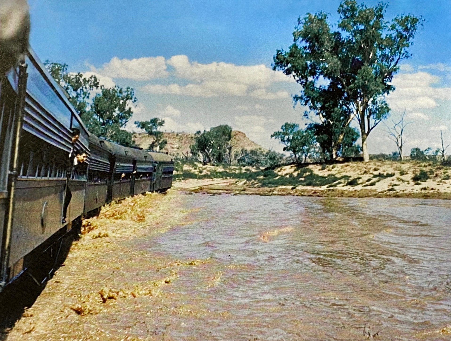 "The Ghan" train crossing the flooded Finke River, Northern Territory, Australia ca 13 Feb 1953. In early 1953, a tropical low-pressure system disrupted the Central Australia Railway for six weeks. The first Ghan service after the "big wet" left Port Augusta on 9 February and took five days to reach its destination, Alice Springs. The train had to stop at several places to allow sand to be removed from the track and there was a two-day delay at the Finke River while the locomotive crew waited for the water level to drop. Then the train proceeded slowly across when the water was still 250 mm (10 inches) above the rails, which because of previous bridge washaways had been laid across the river bed. When diesel-electric locomotives were used, it was no longer possible to run where water covered the rails because low-slung electric traction motors in the bogies would be affected.