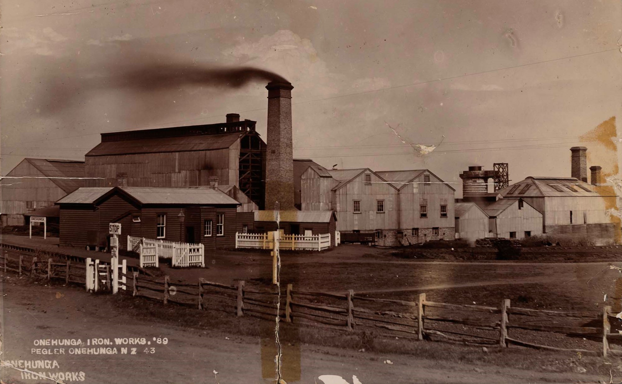 This description is taken from the Auckland Museum website:
"Image showing a large factory complex comprised of a number of tin and small weatherboard buildings. A large chimney stack made of bricks rises in the centre, with black smoke coming out of the top. A simple wooden fence runs along the length of the property, with a white sign reading 'STOP' at the gate. To the left of the image, behind a dark weatherboard building, is another white sign reading 'ONEHUNGA'. To the right of the image, behind a large metal building, is a large blast furnace and two more large brick chimneys."
The date of 1889 is based on the following; the number " '89 " on the handwritten caption on the image; the fact that the plant is in production as evidenced by the smoke from the chimney (making it earlier than August 1895), and the fact that the blast furnace is shown (it was under construction in July 1889 and in production in June 1890).
This photograph is from the Ellen Louise McLeod collection of photographs of the Auckland Museum. The Museum's website state that there are No Known Copyright Restrictions.