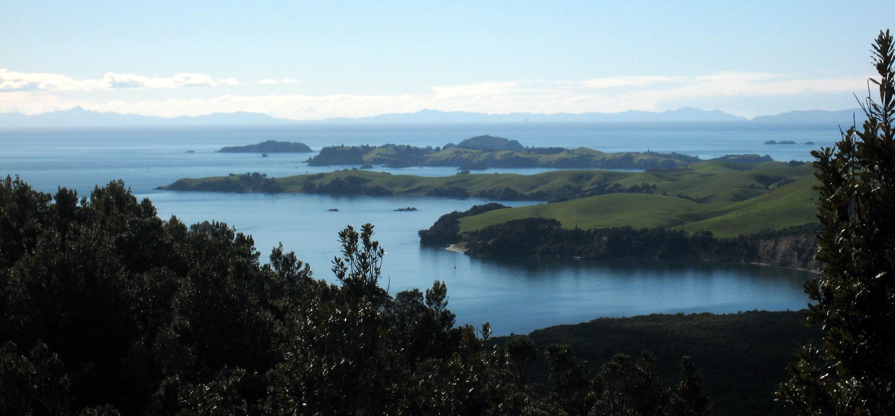 View from near the summit of Rangitoto Island, Auckland, New Zealand, looking out over Motutapu, Rakino and the Noises. The closest two grassy headlands are part of Motutapu Island. Rakino Island, almost as grassy, is a little more distant. Bush-covered Otata Island, the largest of the Noises group, peers over Rakino, and Motuhoropapa Island (the second largest) lies to Rakino's left. Great Barrier Island is visible along the horizon, with the tip of the Coromandel Peninsula appearing in the far right.
