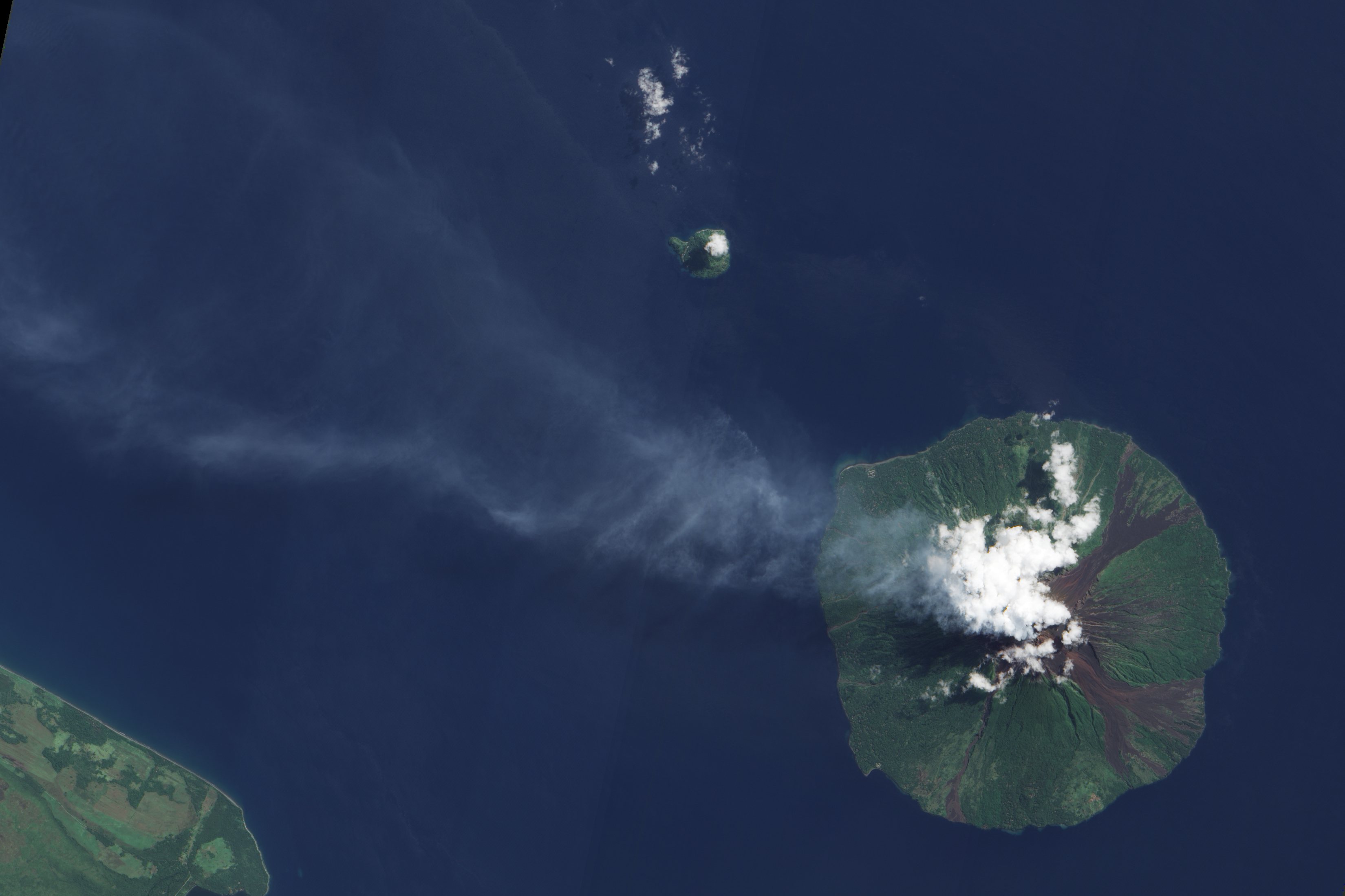Bright white clouds hover over the volcano’s summit. Clouds often collect over peaks, but these clouds could result from water vapour released by the volcano. Slightly darker in colour, a pale blue-grey plume blows west-north-west from the summit and over the Bismarck Sea. Just 10 kilometres across, Manam is a stratovolcano composed of alternating layers of ash, lava, and rocks from prior eruptions. The circular island sports four radial valleys spaced roughly 90 degrees apart, and these valleys have historically channelled lava and pyroclastic flows—composed of hot rock, gas and ash—sometimes all the way to the coast. Evidence of earlier volcanic activity appears as rivulets of rock that interrupt the vegetation coating much of the island. Although clouds hide the summit in this image, the summit is known to support two craters devoid of vegetation.