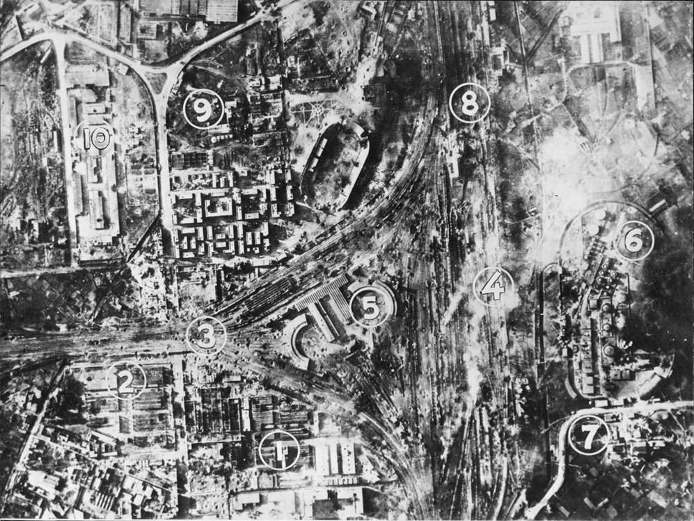 This photograph, taken from a reconnaissance plane, shows some of the damage to the Italian port of Naples caused by 500 Allied bombers in the greatest combined air attack up to that time by Allied air squadrons operating against the Axis from bases in North Africa. In the attack, launched in early morning darkness by R.A.F. Wellingtons and continued through one and a half hours of daylight by U.S.A.A.F. bombers, crows observed tremendous explosions as the Royal Arsenal was blown up with destruction or damage to all its buildings. All buildings of the torpedo works and of an ammunition train was blasted to bits. Further damage is indicated by other numerals as follows: (3) railroad tracks torn up, with craters clearly visible; (4) freight and passenger trains damaged, destroyed and burning in the central area of the marshalling yards; (5) an engine roundhouse and carriage and repair sheds heavily damaged, with countless hits on tracks and cool dumps; (6) oil tanks destroyed, with fires still burning furiously; (7) a factory destroyed; (8) tracks broken and large buildings destroyed at the north exit from the Naples marshalling yards, and (9) an oil factory heavily damaged.