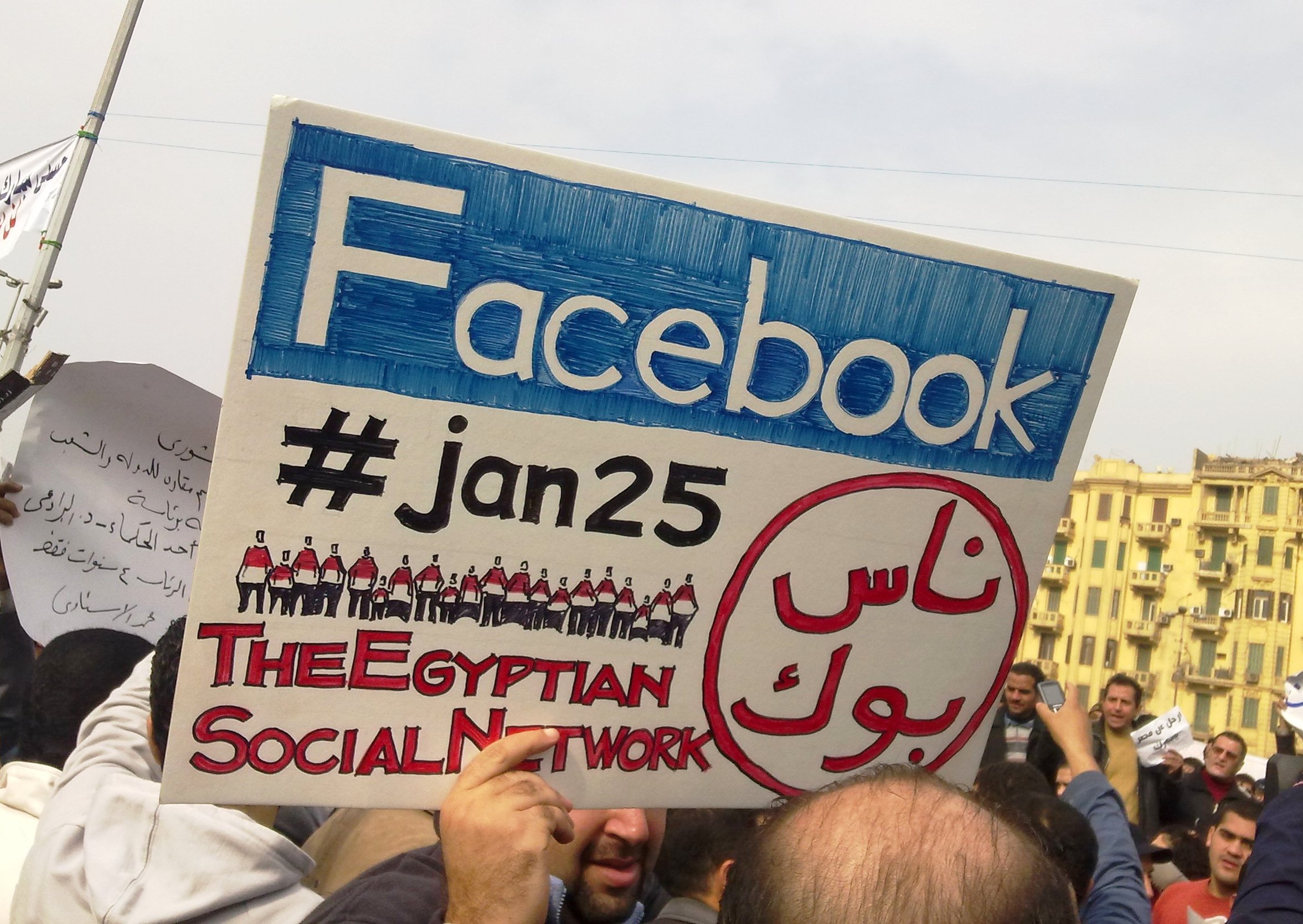 a man during the 2011 Egyptian protests carrying a card saying "Facebook, #jan25, The Egyptian Social Network".