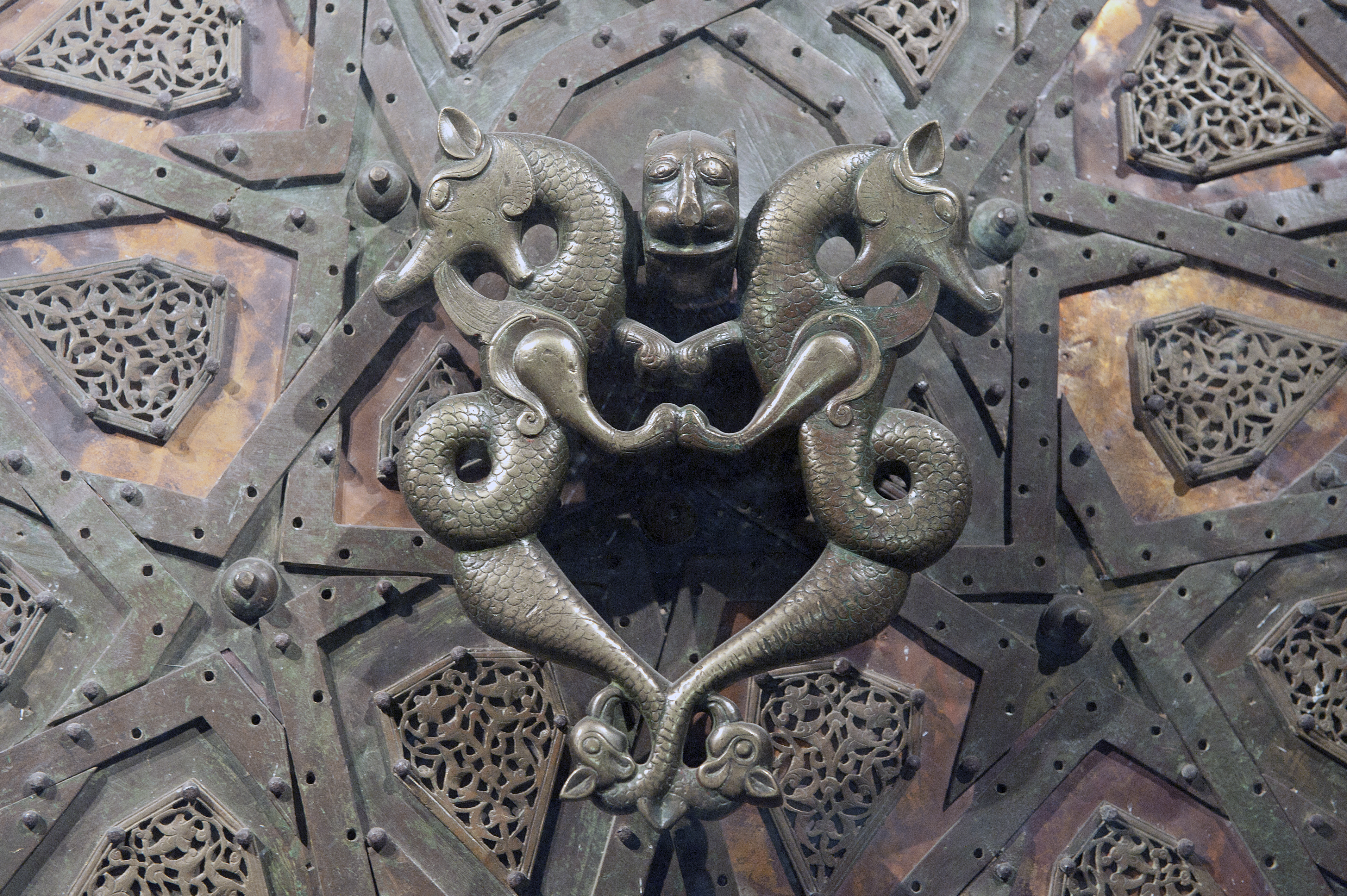 The double doors of the Cizre mosque (built 1155-1160 by the Atabegs of Mosul) measure 300 x 112 cm. and consist of a timber frame plated with beaten copper sheets attached to the wood by nails. They are elaborately decorated with brass rods forming an interlocking design of twelve-pointed stars, in the spaces between which are plaques with openwork designs of scrolls with rumi-palmette motifs. A text in a band across the tops of the doors is supposed to be a later addition. One of the two identical doorknobs was stolen in 1969 and taken to the David Collection in Copenhagen. The lion holding it remained in place. A double dragon is frequently found on Artukid coins. They are often combined with lions, together they protect and represent sun and moon. (From information on site)