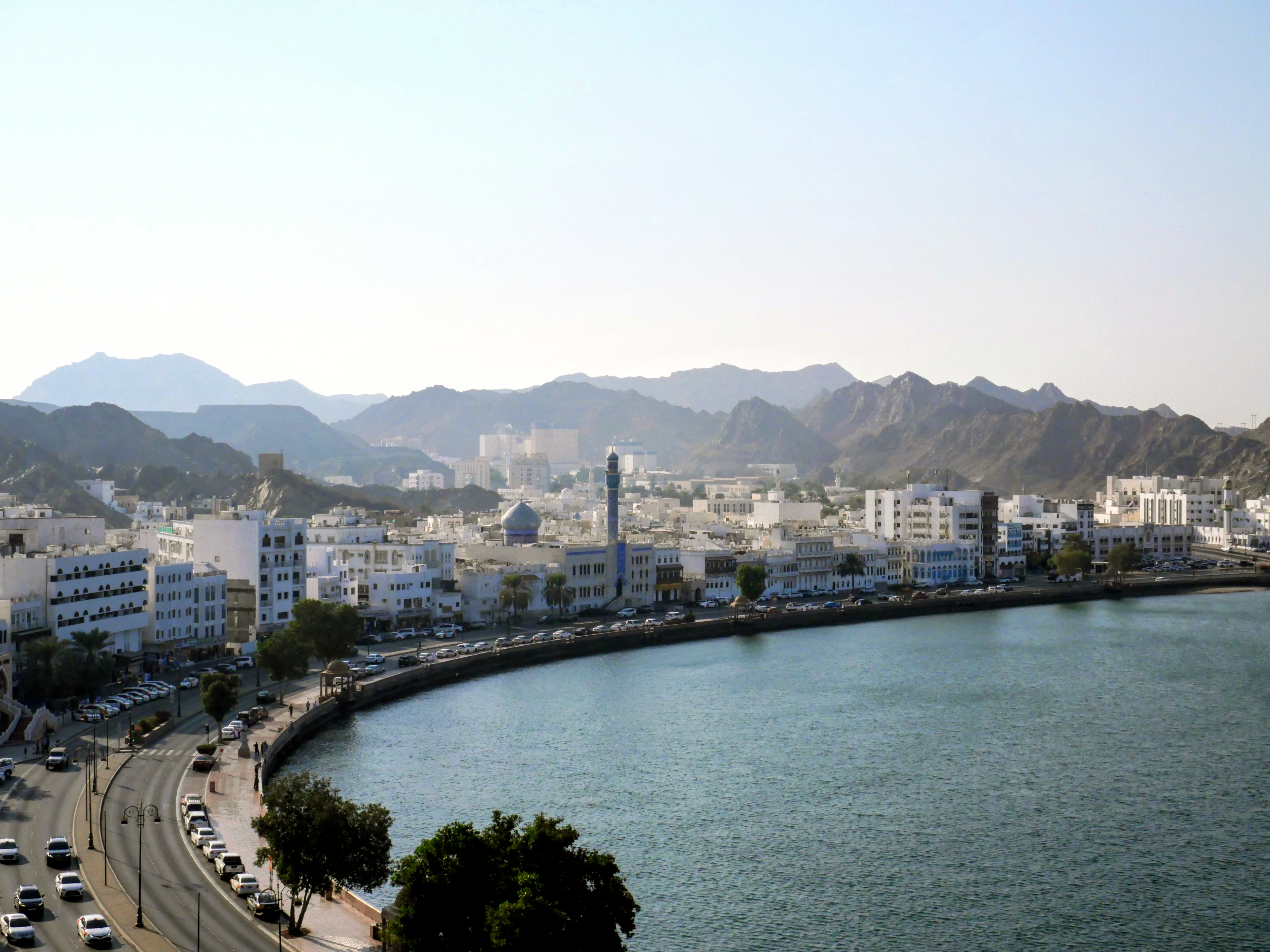 A panoramic view of the Muttrah Corniche and harbor in Muscat, Oman, featuring the white buildings of the city curving along the waterfront, backed by the dramatic, arid mountains of the Al Hajar range.