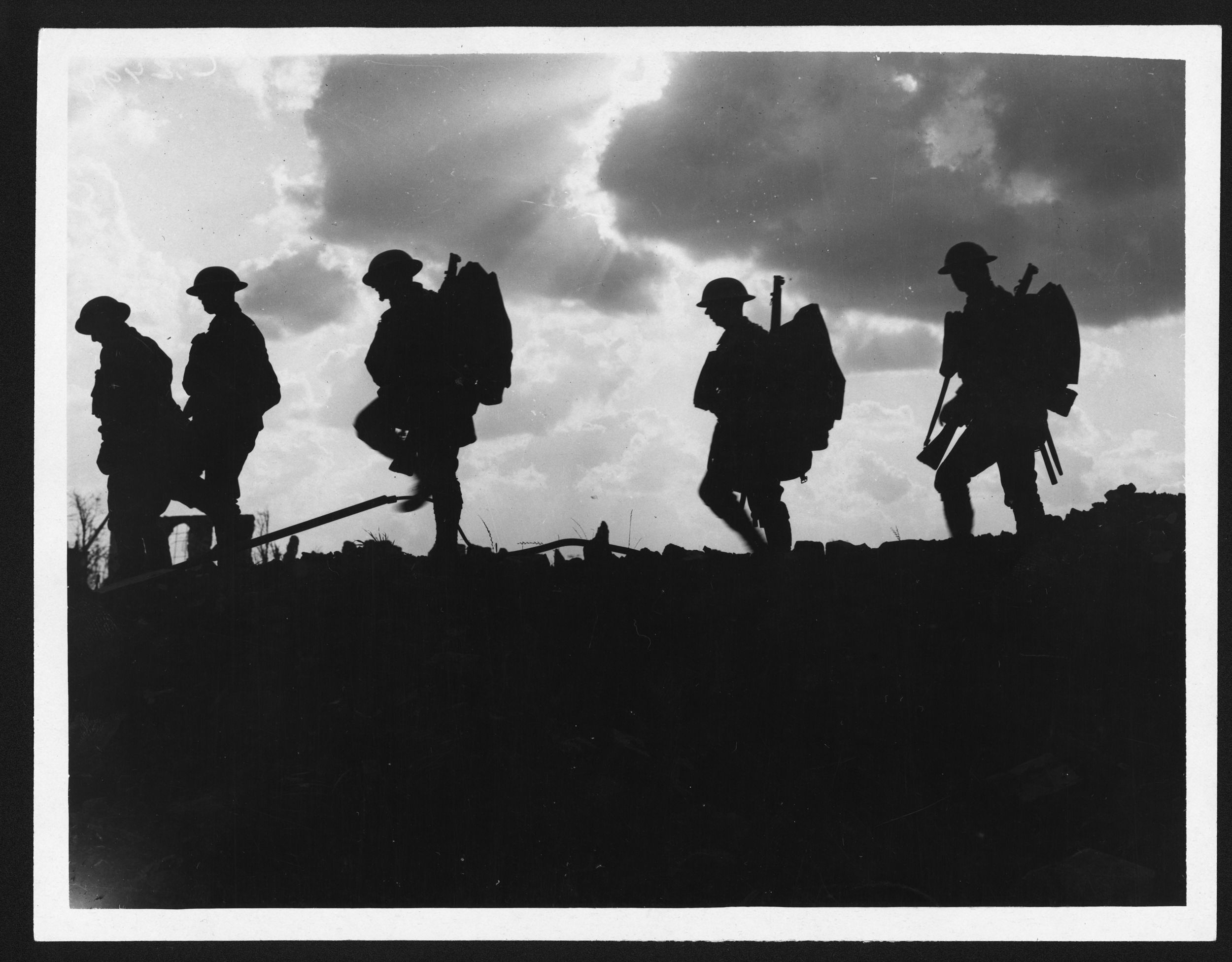 Five soldiers silhouetted against the sky. Rays of sun burst through dark clouds to create a dramatic and atmospheric shot. They are all wearing steel helmets, and three of them are clearly carrying rifles and backpacks.Original reads: 'BATTLE OF BROODSEYNDE [sic] RIDGE. - TROOPS MOVING UP AT EVENTIDE. MEN OF A YORKSHIRE REGIMENT ON THE MARCH.'The Imperial War Museum caption (Q 2978) gives "A silhouetted file of men of the 8th Battalion, East Yorkshire Regiment going up to the line near Frezenberg.")