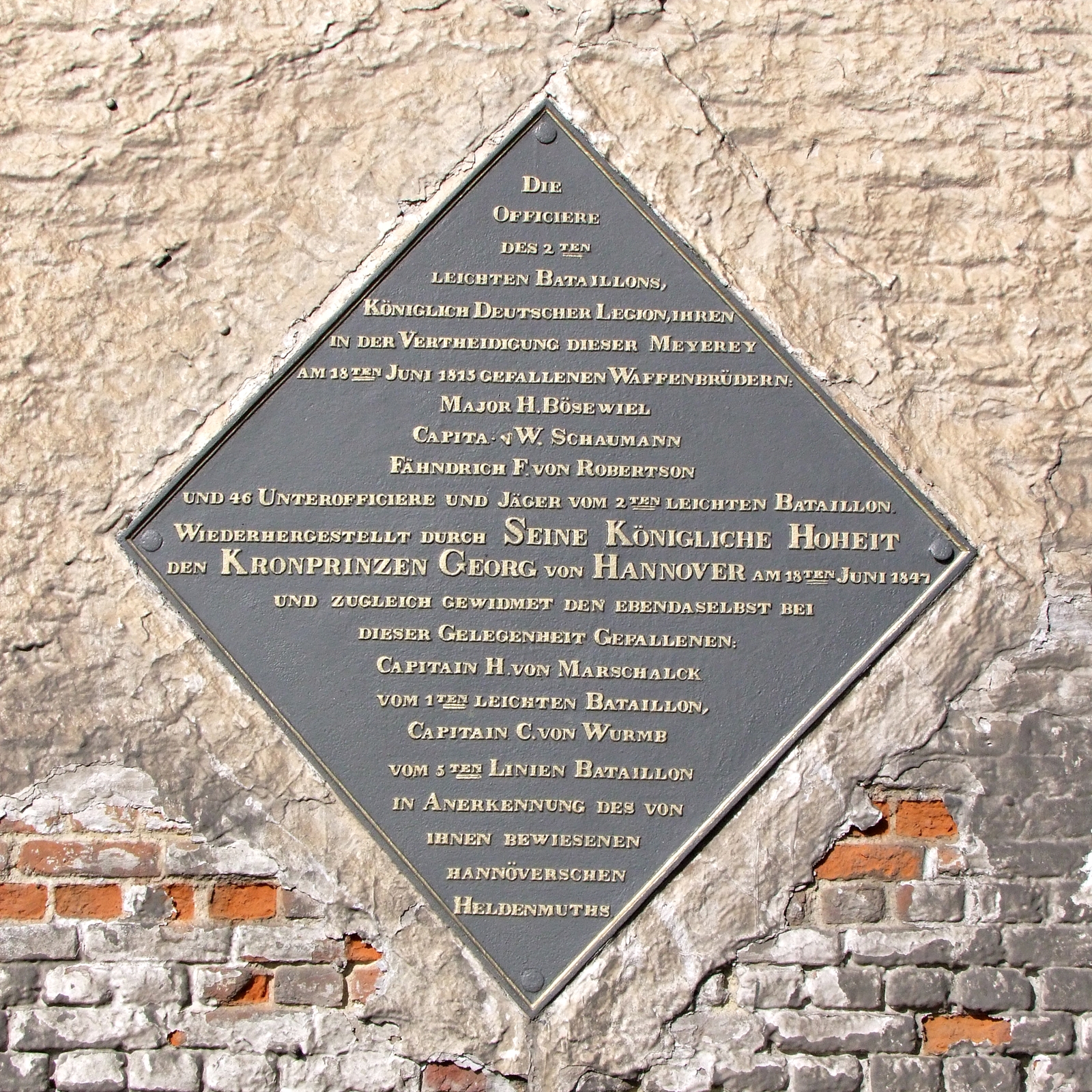 Plaque imbedded in 1847 in the eastern gable of the La Haye Sainte main building, overlooking the road N.5, replacing a previous marble tablet presumably affixed in 1818, along with the construction of the monument to the K.G.L. : ►(File:18 June 1815 – Victory at Waterloo – Hanoverian Monument, The South Face.jpg).  

About the plaque. 
En mémoire de la défense de la Haie-Sainte, le corps des officiers Anglo-Hanovriens du deuxième bataillon léger de la légion royale allemande fit encastrer, dans le pignon de la ferme vers la route, une plaque de marbre blanc, où on rappelait la mort glorieuse de plusieurs officiers de ce bataillon ; en 1847, le prince George, actuellement roi de Hanovre, l'a fait remplacer par une plaque de fer carrée et posée en losange.  On y lit, en lettres saillantes et dorées :  (...).
La ferme a été rebâtie et est, comme jadis, bornée au sud par un verger.  Les champs voisins, où le carnage a été affreux, constituent en réalité une immense nécropole.  Une seule fosse, pratiquée dans la vallon, vers l'est, a reçu les corps de 4,000 soldats et d'un grand nombre de chevaux.

Tarlier, Charles ; Wauters, Alphonse, Géographie et histoire des communes belges, vol. 1, Novembre 1869, 78.


TRANSLATION
In memory of the defense of La Haie-Sainte, the corps of Anglo-Hanoverian officers of the second light battalion of the royal German legion had a white marble tablet embedded in the gable of the farm towards the road, where the glorious death of several officers of this battalion was recalled; in 1847, the prince George, currently King of Hanover, had it replaced by a square iron plate laid in a diamond pattern. It reads, in protruding and gilded letters: (...).


The farm has been rebuilt and is, as in the past, bounded to the south by an orchard. The neighbouring fields, where the carnage was horrendous, are in reality a huge necropolis. A single pit, made in the valley to the east, received the bodies of 4,000 soldiers and a large number of horses.