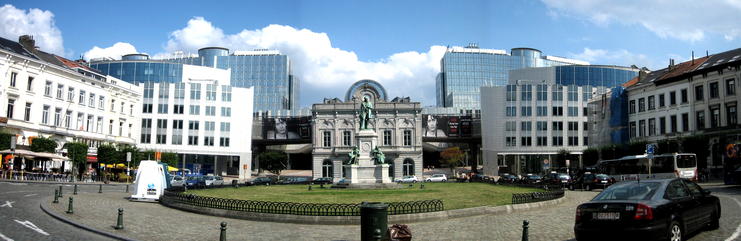 Panorama of the Place du Luxembourg, European Quarter of Brussels (Belgium). View of the European Parliament (western side), including converted station entrance in front) with statue of John Cockerill in foreground. Restaurants and bus stops on pavements each side.