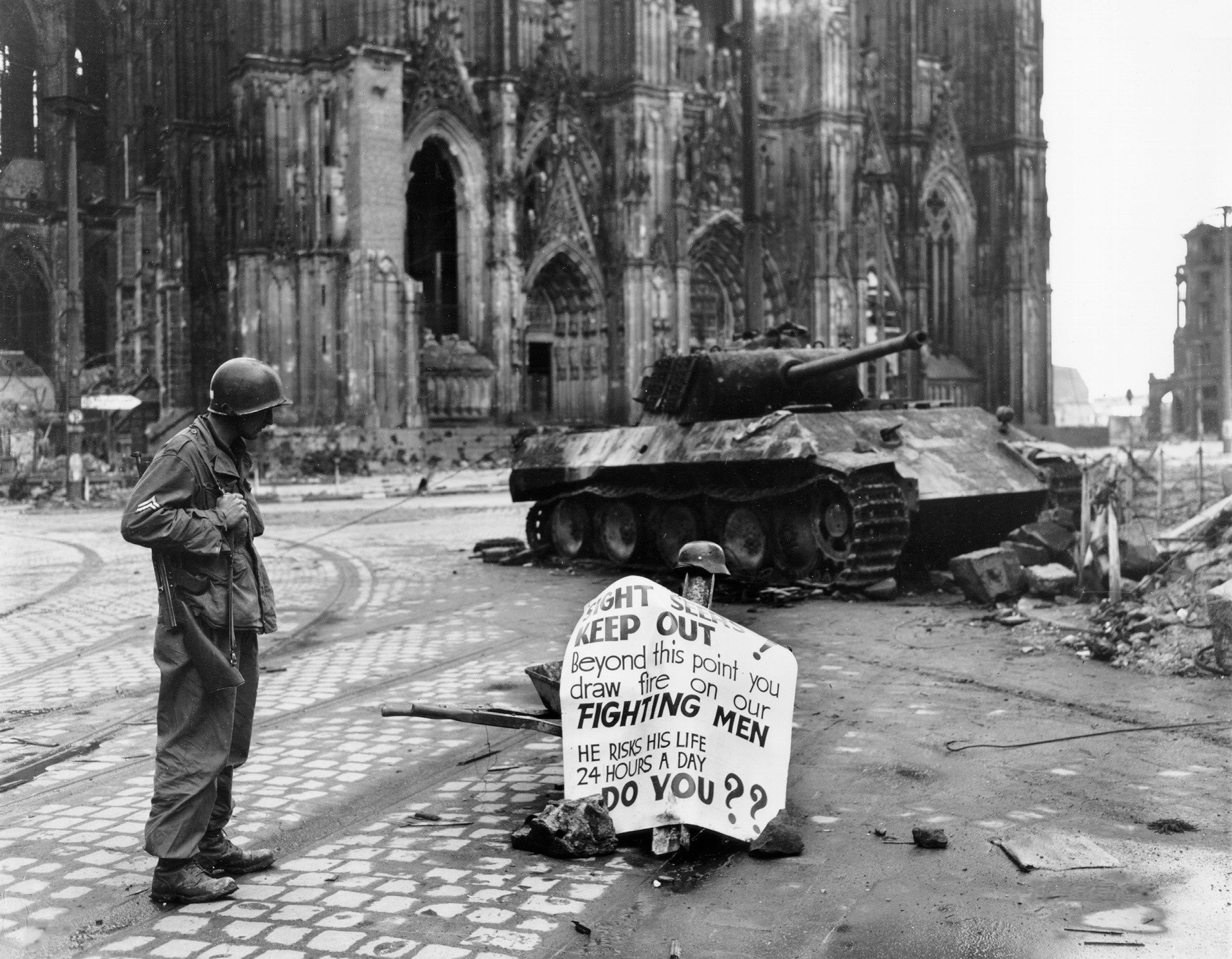 Original caption: Cologne, Germany - Cpl. Luther E. Boger, Concord, N.C., skytrooper, reads a warning sign in the street. This street leads to the Rhine River and is under observation of the Germans who occupy a stronghold there. Cpl. Boger is with the 82nd Airborne Division. 4 April 1945. The German tank is burnt out and the torsion-bars have been destroyed by the immense heat of the fire.

Allied troops captured the western part of Cologne on 6th-7th March 1945. The German army still held the eastern shore of the Rhine and attacked the Allies with artillery. The rest of Cologne was captured between 12th and 15th April 1945. Until the 16th April a strip of about 500m along the shore had been declared as a restricted area and the cathedral was just within this zone.

Text on sign reads:

SIGHT SEERS
KEEP OUT!
Beyond this point you
draw fire on our
FIGHTING MEN
HE RISKS HIS LIFE
24 HOURS A DAY
DO YOU??