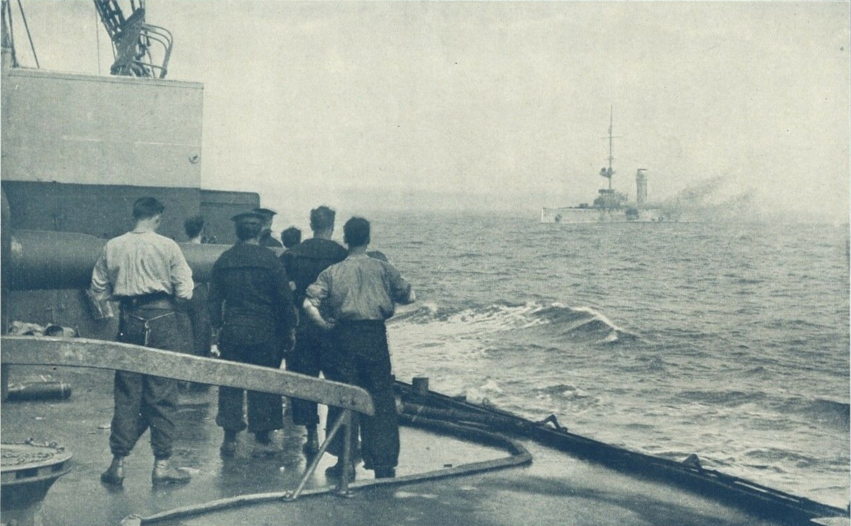 Titleː A Remarkable Camera Record of the Sinking of the German Cruiser Mainz off Heligoland August 28th 1914. A photograph taken from the deck of a British cruiser just before the Mainz sank, In the foreground British sailors watch the Mainz on fire.
The text indicates the photo was taken from the deck of one of the British cruisers present, and shows a British gun crew standing by their 6 inch gun. The Mainz is seen on fire, minus two after funnels and a mast, shortly before heeling over and sinking.