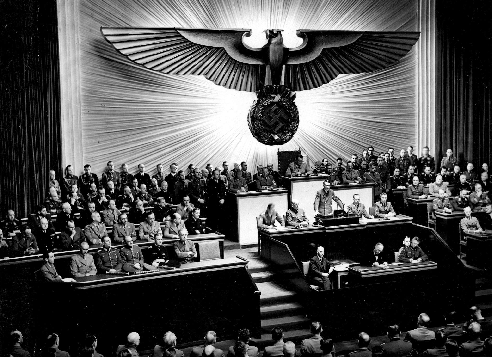 For documentary purposes the German Federal Archive often retained the original image captions, which may be erroneous, biased, obsolete or politically extreme. Berlin, Reichstagssitzung, Rede Adolf Hitler
Die vernichtende Rede des Führers über Roosevelt.
Am Donnerstag nachmittag hielt der Führer vor den Männern des Deutschen Reichstags die grosse und mit fieberhafter Spannung erwartete Rede zu dem von dem Kriegshetzer Roosevelt heraufbeschworenen Krieg im Pazifik. 

Auf der Regierungsbank (von rechts nach links) sieht man
den Führer, neben ihm
Reichsaussenminister v. Ribbentrop,
Grossadmiral Reader,
Generalfeldmarschall v. Brauchitsch,
Generalfeldmarschall Keitel und die
Reichsminister Dr. Frick und
Dr. Goebbels.
- In der zweiten Reihe (von rechts):
Die Reichsminister Graf Schwerin-Krosigk,
Funk,
Darré,
Rust,
Kerrl,
Dr. Frank,
Dr. Dorpmüller,
Dr. Seyss-Inquart und
Dr. Todt.
- Dahinter (von rechts):
Reichsminister Rosenberg und die
Staatsminister Dr. Meissner und
Dr. Popitz.
AV 65979
[Berlin, Kroll-Oper.- Reichskanzler Adolf Hitler während seiner Rede vor dem Reichstag zur Kriegserklärung an die Vereinigten Staaten.]
Abgebildete Personen:

Hitler, Adolf: Reichskanzler, Deutschland
Ribbentrop, Joachim von: Außenminister, NSDAP, Deutschland
Frick, Wilhelm  Dr.: NSDAP, MdR, Reichsinnenminister, 1924 Hitlerputsch-Prozeß vor dem Volksgericht München,  1946 hingerichtet, Deutschland  (GND 119055201)
Goebbels, Joseph: Reichsminister für Volksaufklärung und Propaganda, Gauleiter Berlin, Deutschland
Schwerin von Krosigk, Lutz Graf: Reichsfinanzminister, Deutschland
Rosenberg, Alfred: Außenpolitisches Amt, Ideologe, Reichsminister Ostgebiete, Deutschland
Göring, Hermann: Reichsmarschall, Oberbefehlshaber der Luftwaffe, Ministerpräsident von Preußen, Deutschland