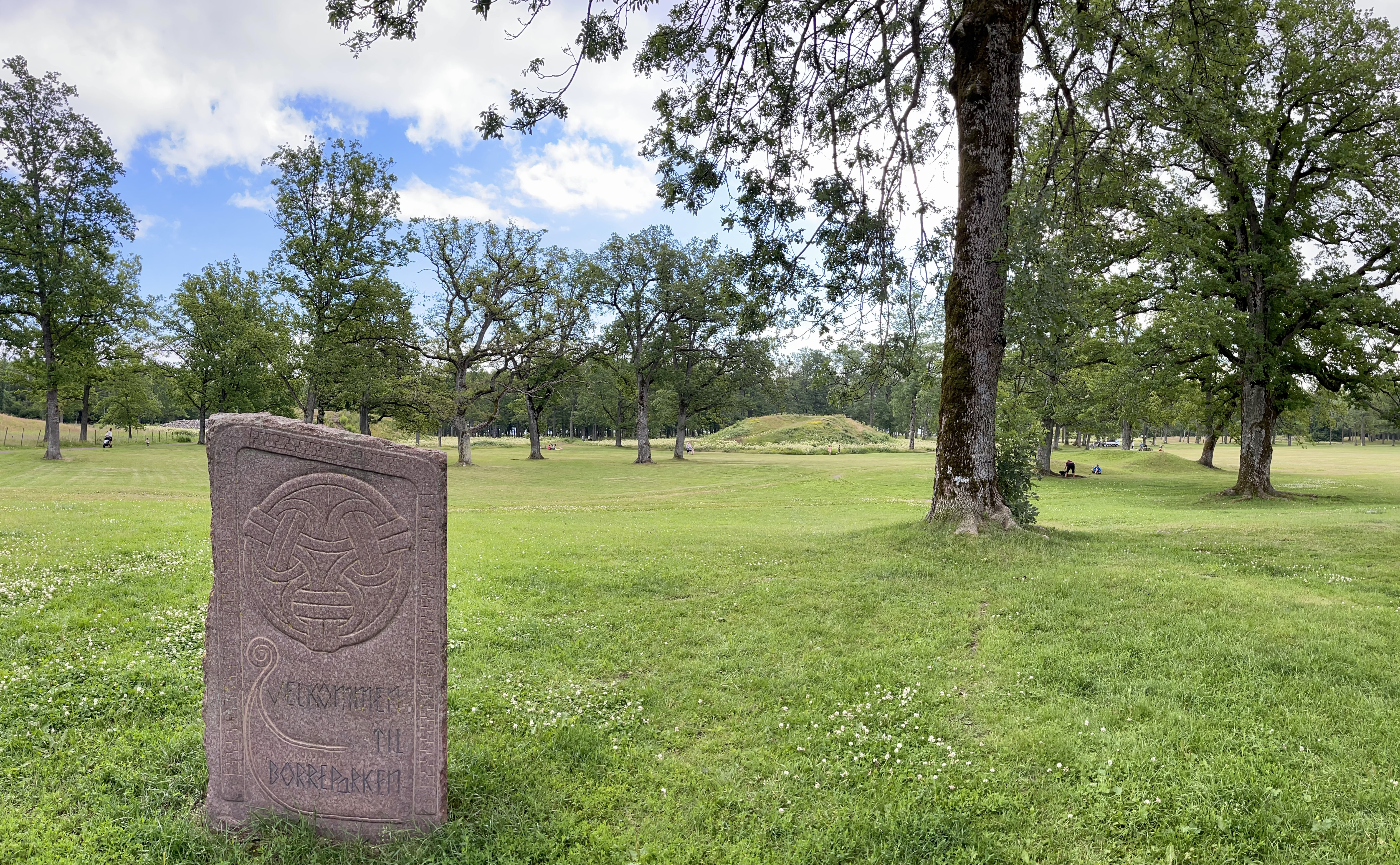 Photo taken at the main entrance/gate to the Borre National Park in the municipality of Horten by the Oslofjorden, Norway. The park contains the Borre mound cemetery (Borre Burial Mounds), a collection of Viking age burial mounds. Close by is the Midgard Vikings Centre (Norwegian: Midgard vikingsenter). Lawn, mounds, trees, summer, stone with the inscription "Welcome to the Borre National Park", etc.