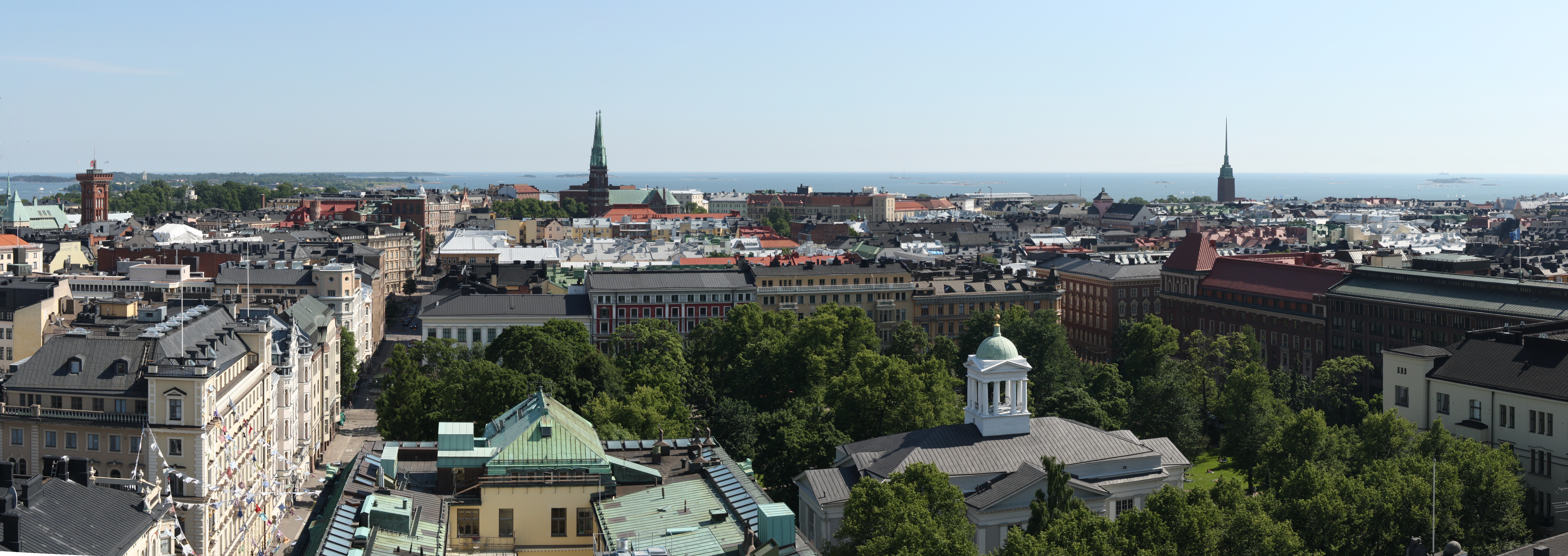 A panoramic view over the southernmost districts of Helsinki from Hotel Torni. The Helsinki Old Church and its surrounding park are seen in the foreground, while the towers of St. John's Church (left of center) and Mikael Agricola Church (right) can be seen in the middle distance, backdropped by the Gulf of Finland.