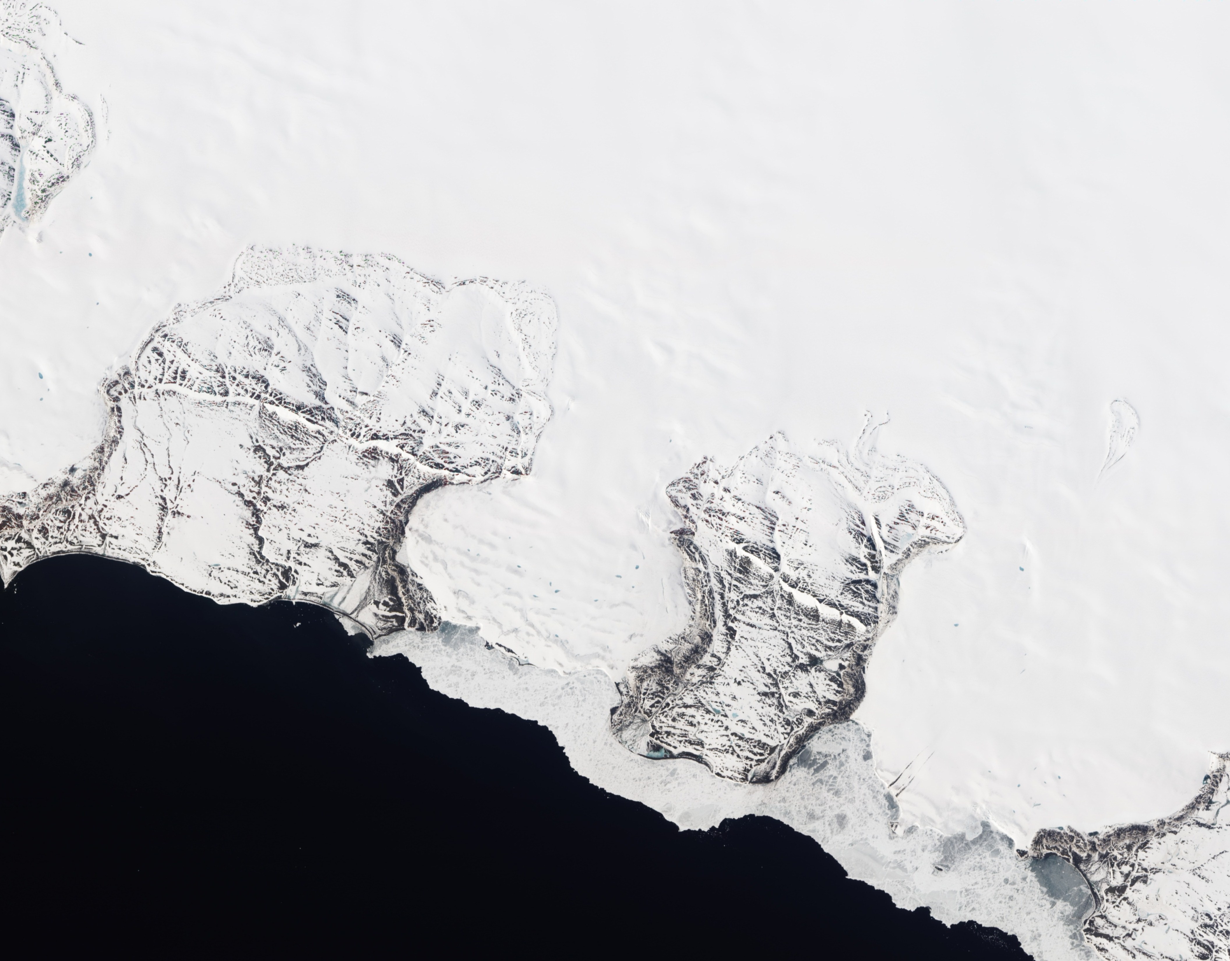 Natural-colour image of Roze Glacier fringed by sea ice. The image is rotated and north is toward the upper right. The glacier pushes slowly seaward between two rocky ridges. Uneven snow cover on the rock surfaces creates a patchwork of brown and white. Some snow also rests on the adjacent sea ice, which appears in shades of white and gray. In the east, a large patch of gray ice immediately offshore may owe its color to a layer of melt water or simply a lack of snow cover. On the glacier itself, isolated pools of melt water form ovals and slivers of blue-gray. Dwarfing the melt ponds, two long parallel stripes extend southward toward the coast. The stripes look like debris along the sides of a relatively fast-flowing ice stream, which may have picked up rocks and dirt from an upslope rock outcrop.